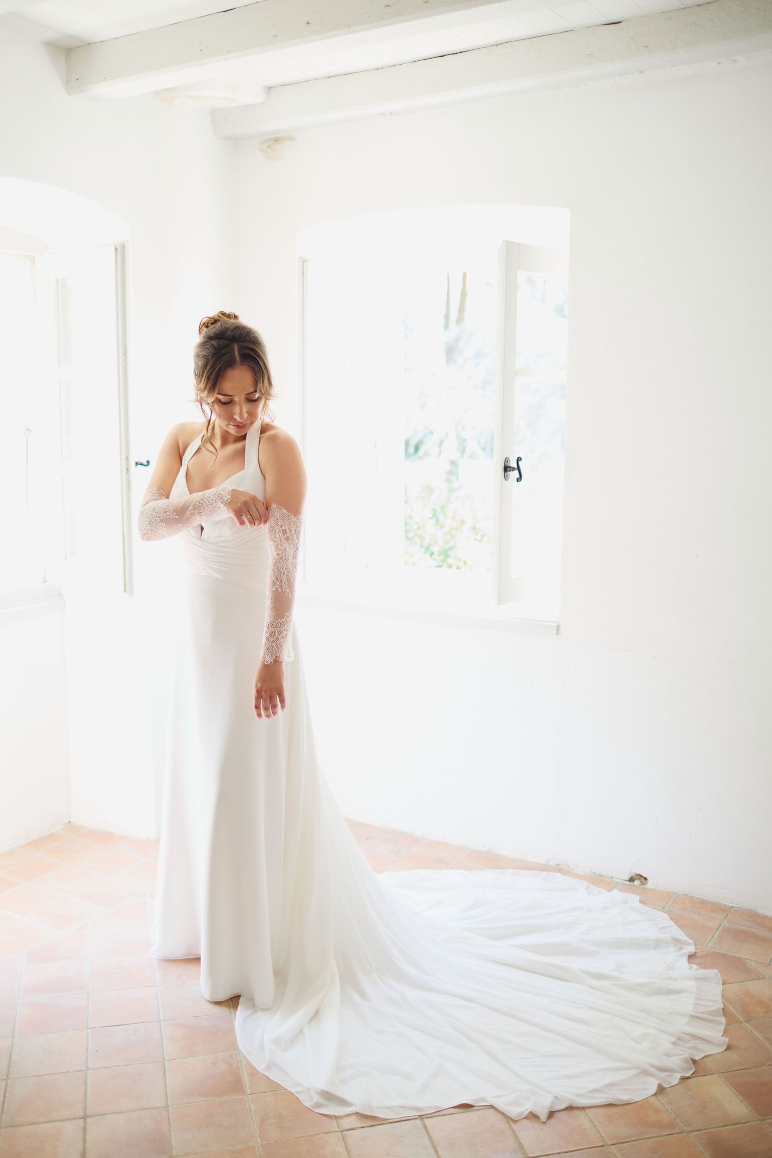 Bride in white wedding dress with lace sleeves and long train standing in sunlit room.