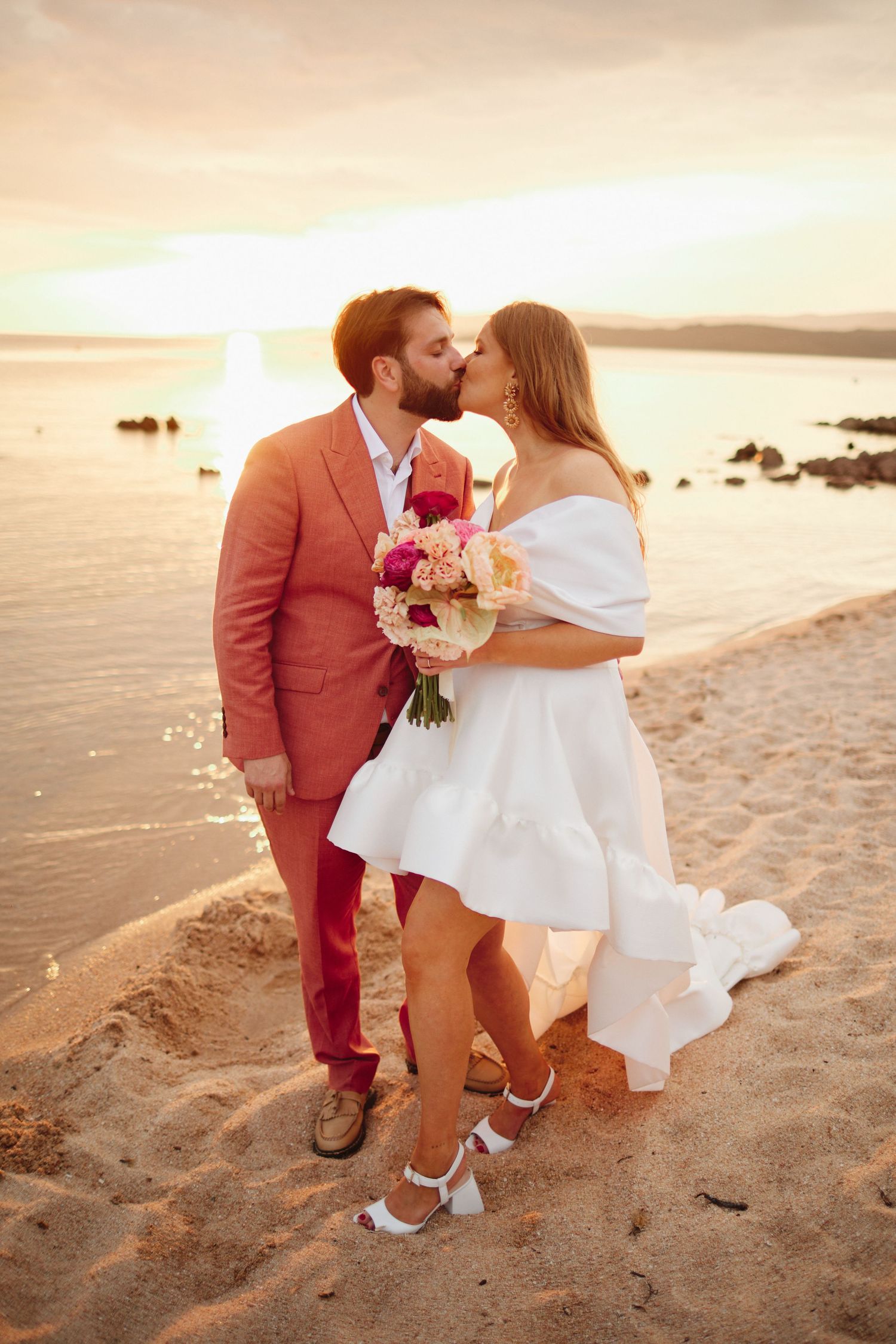 Bride and groom sharing a romantic kiss on a sandy beach during a warm golden sunset.