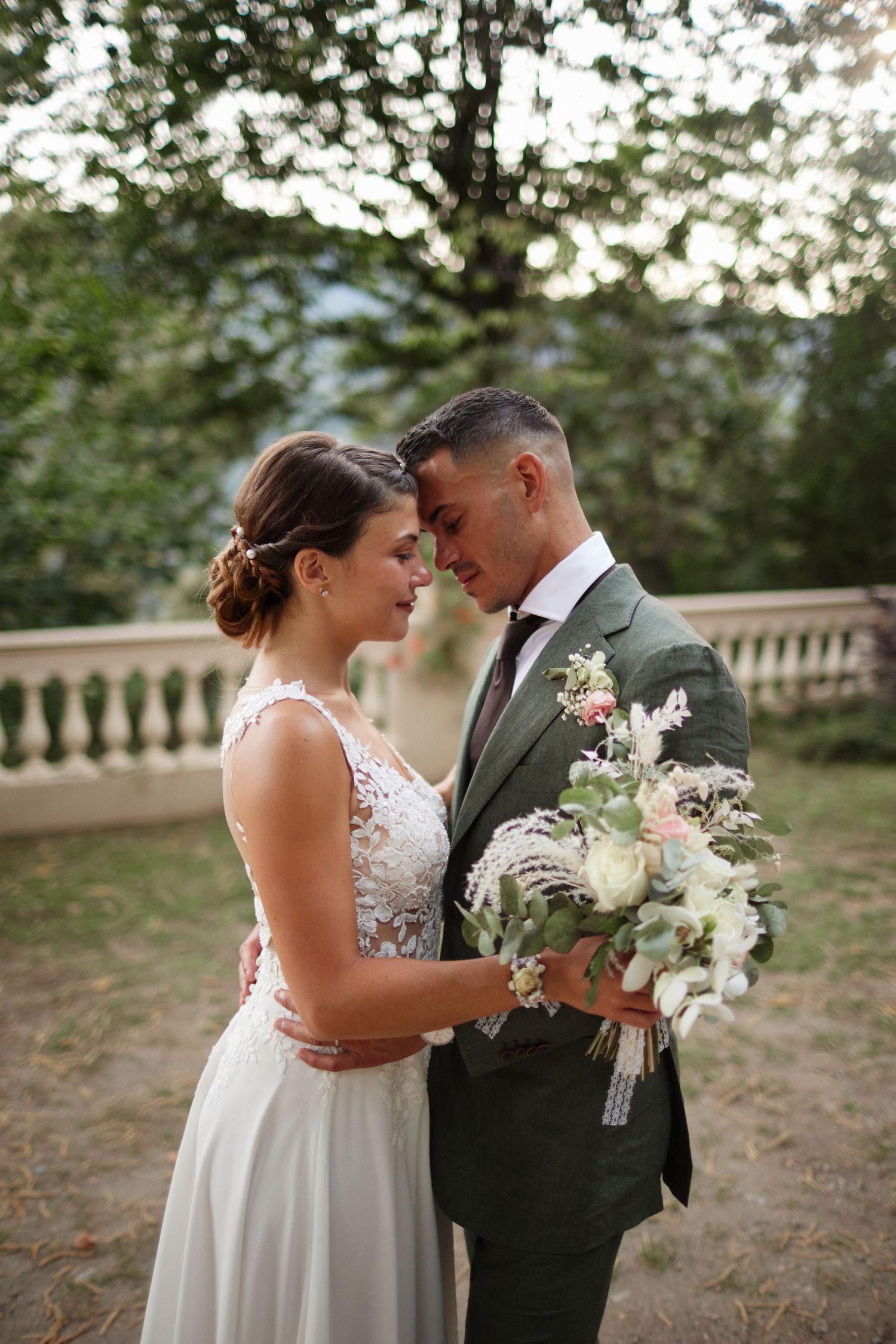 Smiling bride and groom touch foreheads romantically outdoors, groom holding white and pink wedding bouquet.