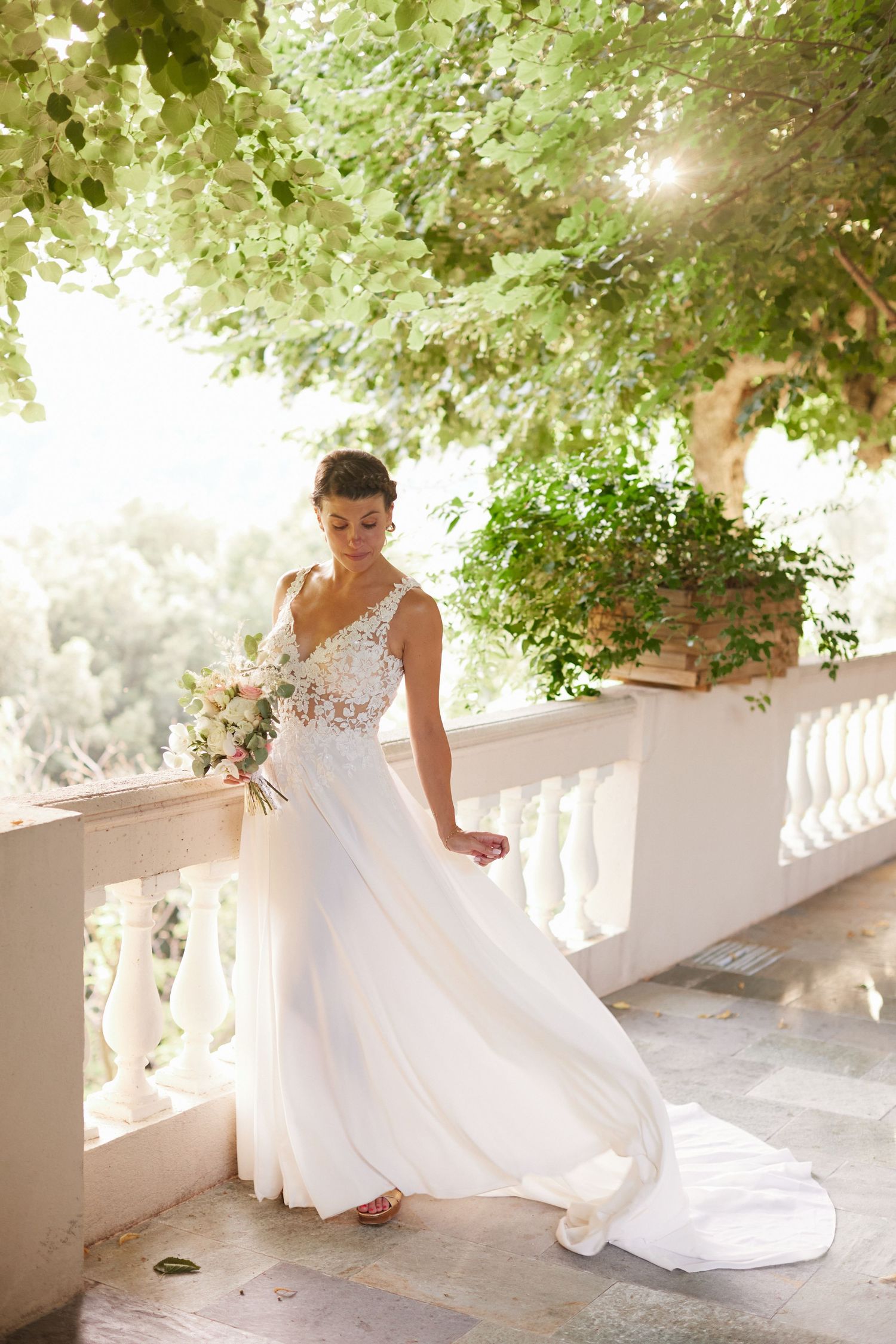 Elegant bride in flowy lace wedding dress holds bouquet on sunlit garden balcony.