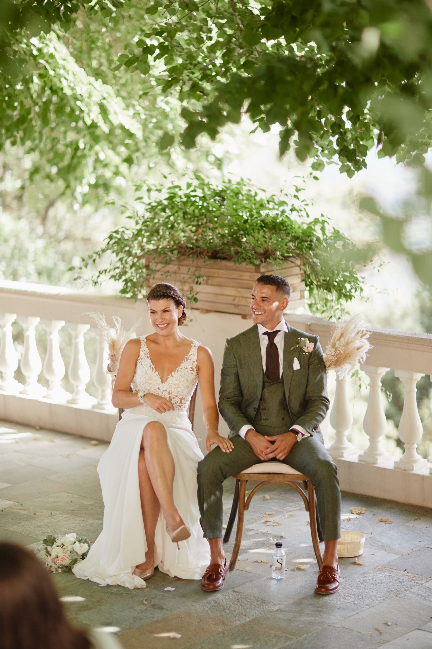 Bride and groom laughing together in wooden chairs during lush outdoor wedding ceremony.