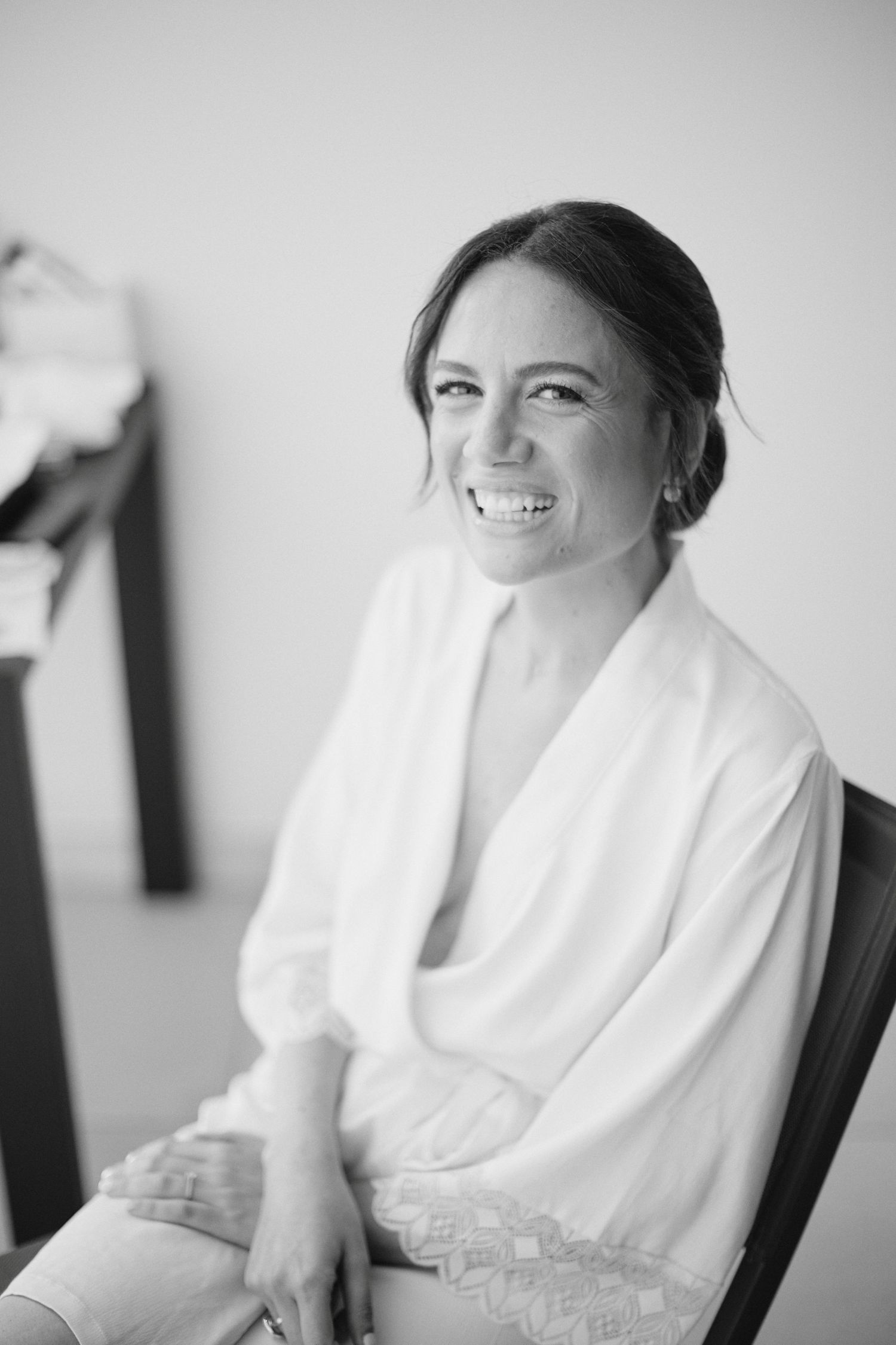 Black and white portrait of radiant bride in white lace robe smiling during wedding morning preparations.