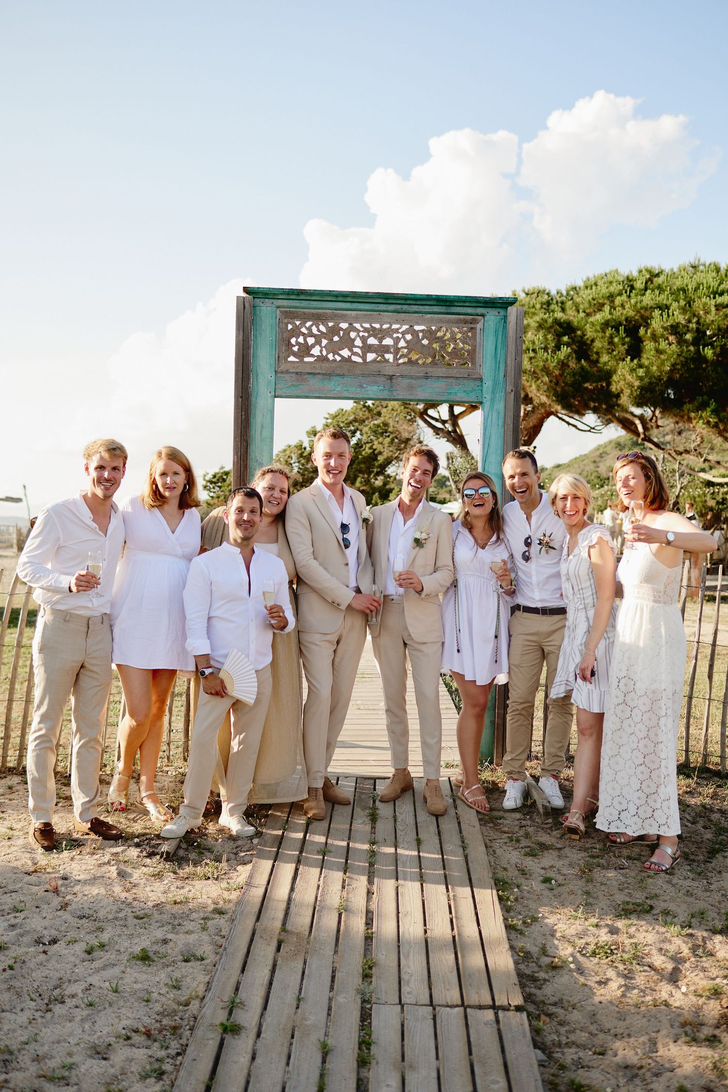 Wedding party group photo on a boardwalk in front of a decorative turquoise frame at a beach venue.