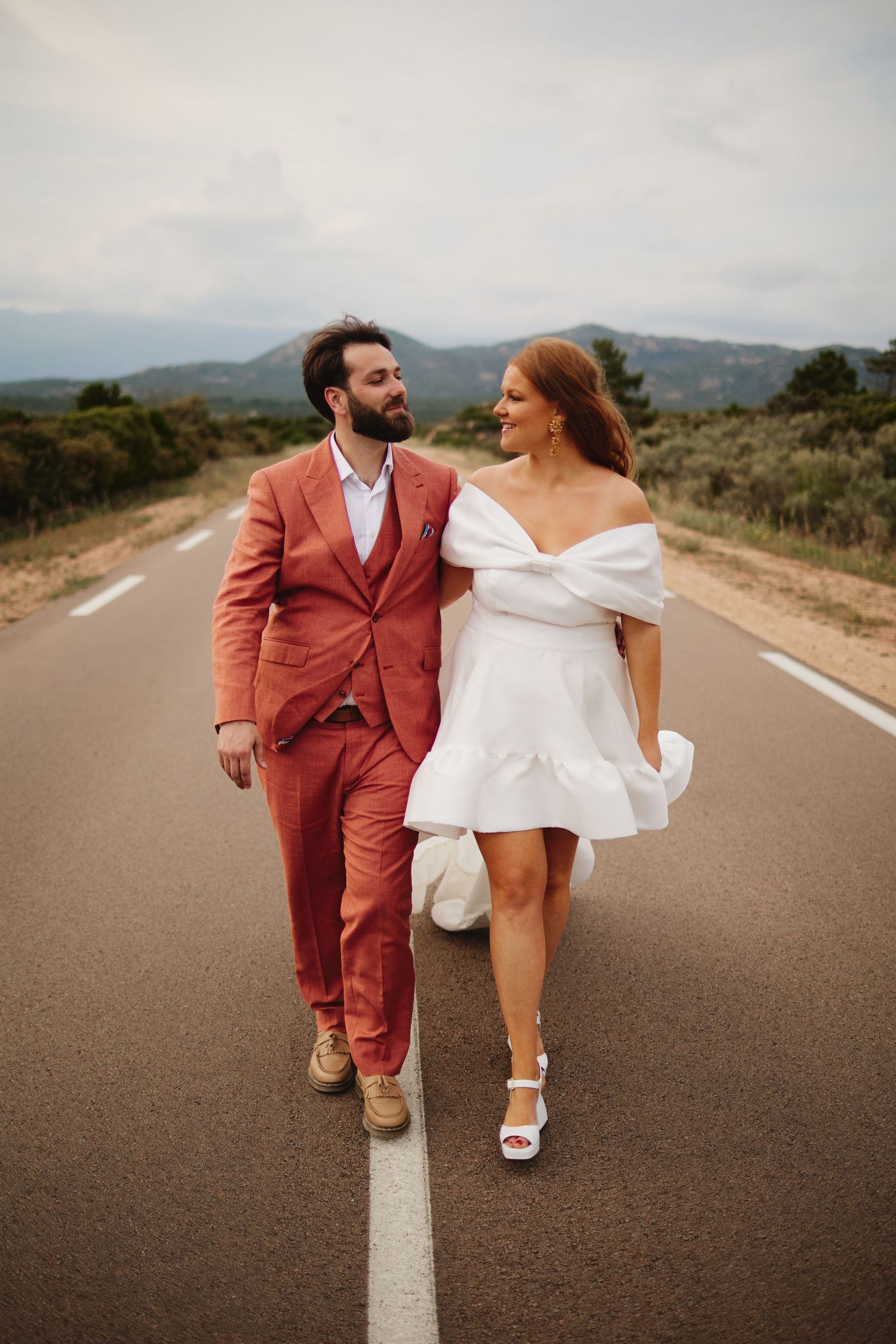 Happy couple in wedding attire walks together down a scenic open road through Mediterranean countryside.