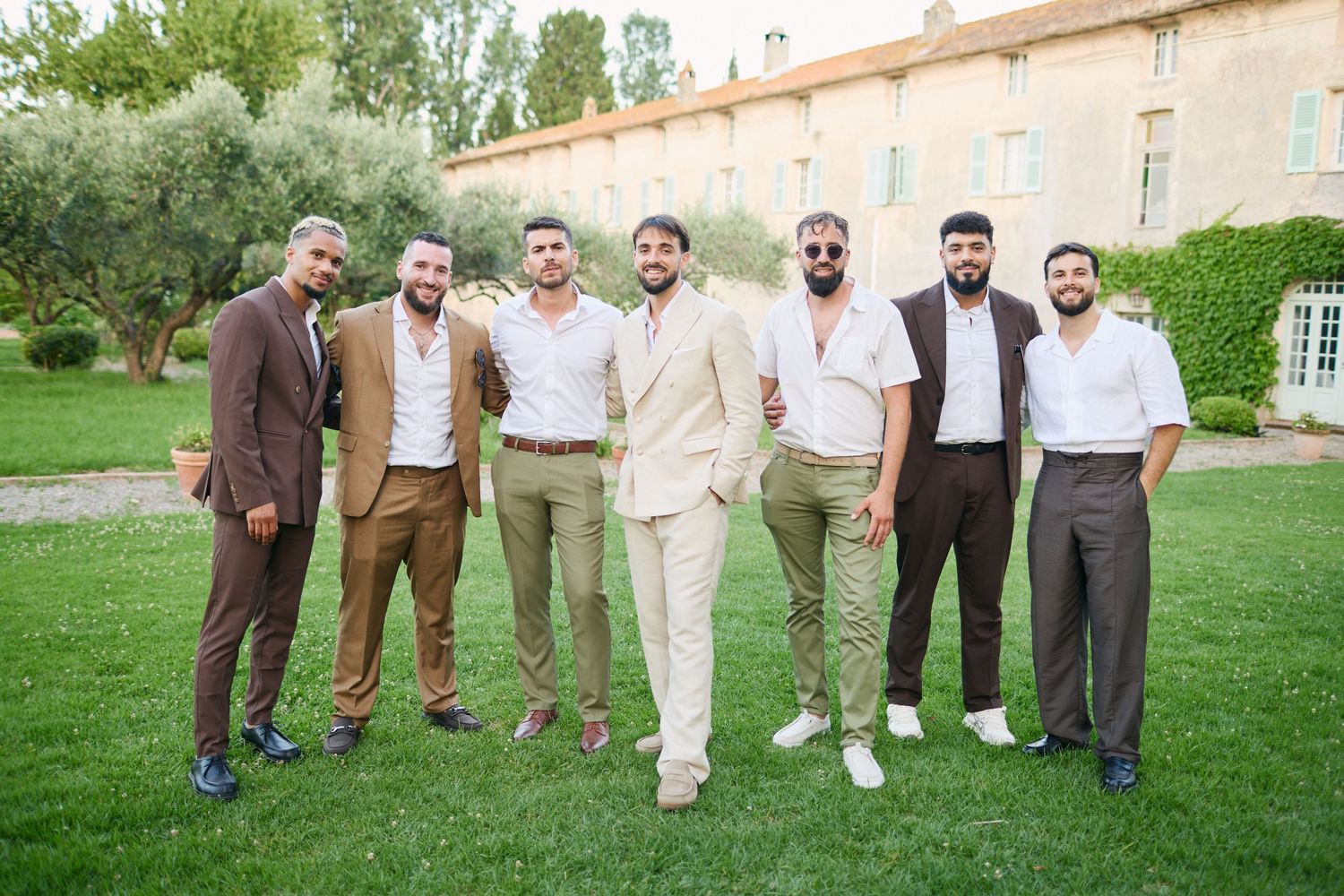 Groom in beige suit posing with six groomsmen in earth tones on green lawn at outdoor wedding.
