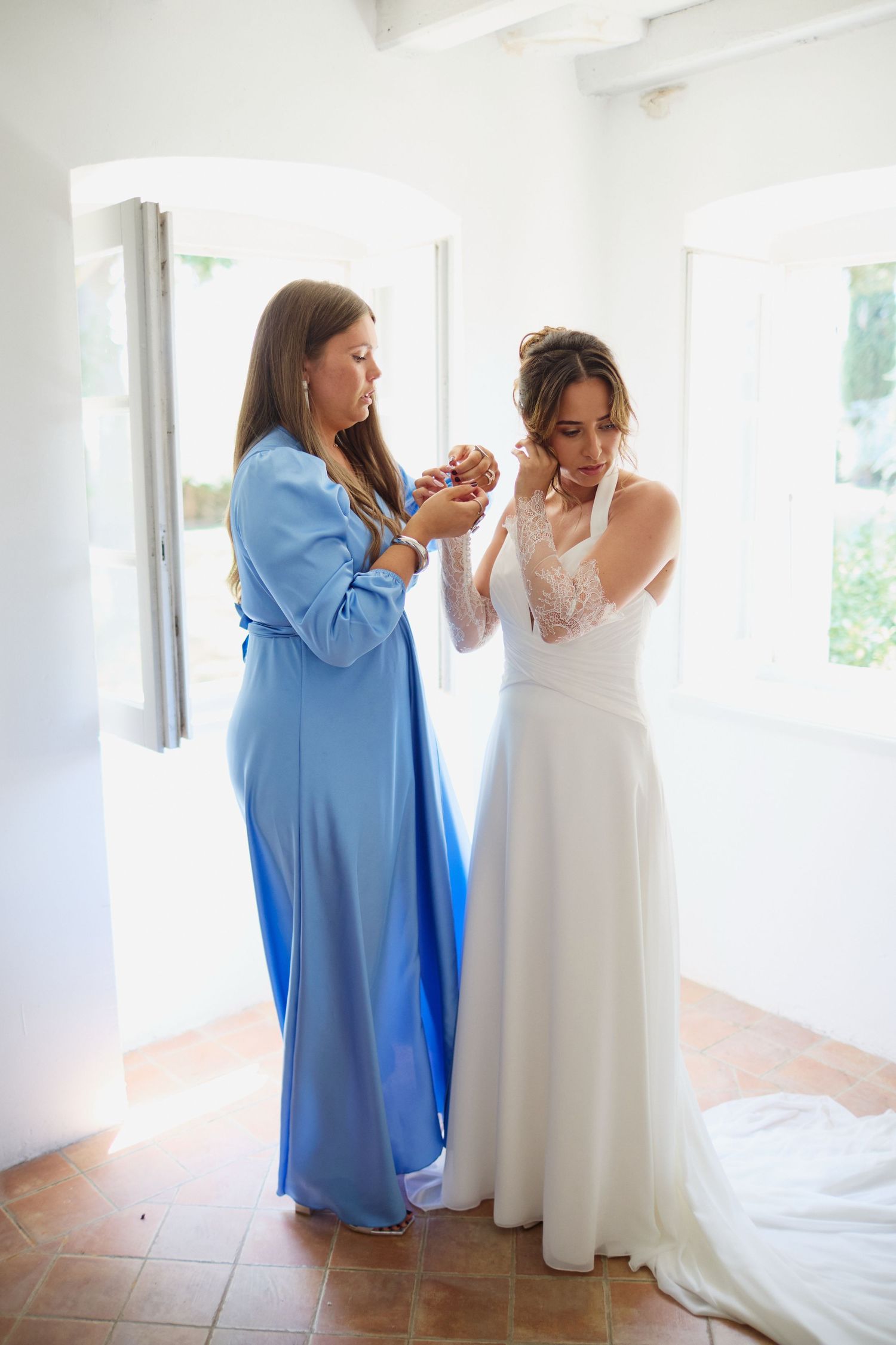 Bridesmaid in blue gown helps bride with earrings while getting ready in bright white room before wedding.