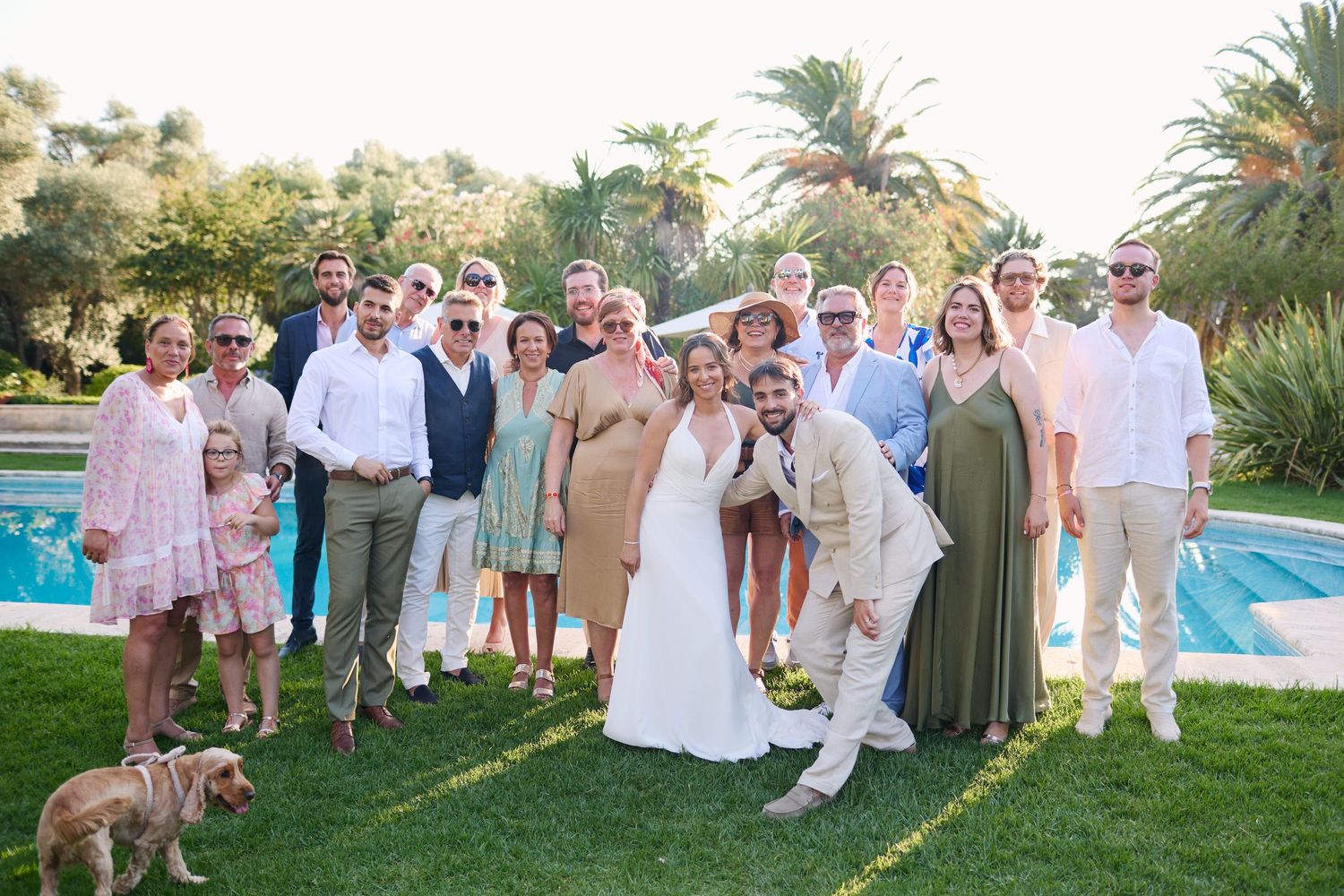 Wedding party and guests pose for group photo beside pool at sunny Mediterranean outdoor wedding reception.