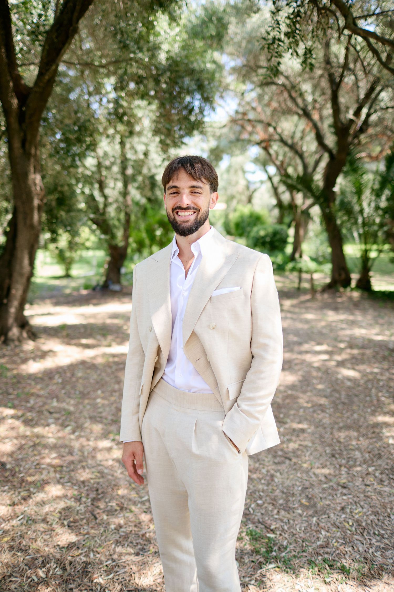 Handsome groom in cream double-breasted suit smiling in a sunlit outdoor garden setting.