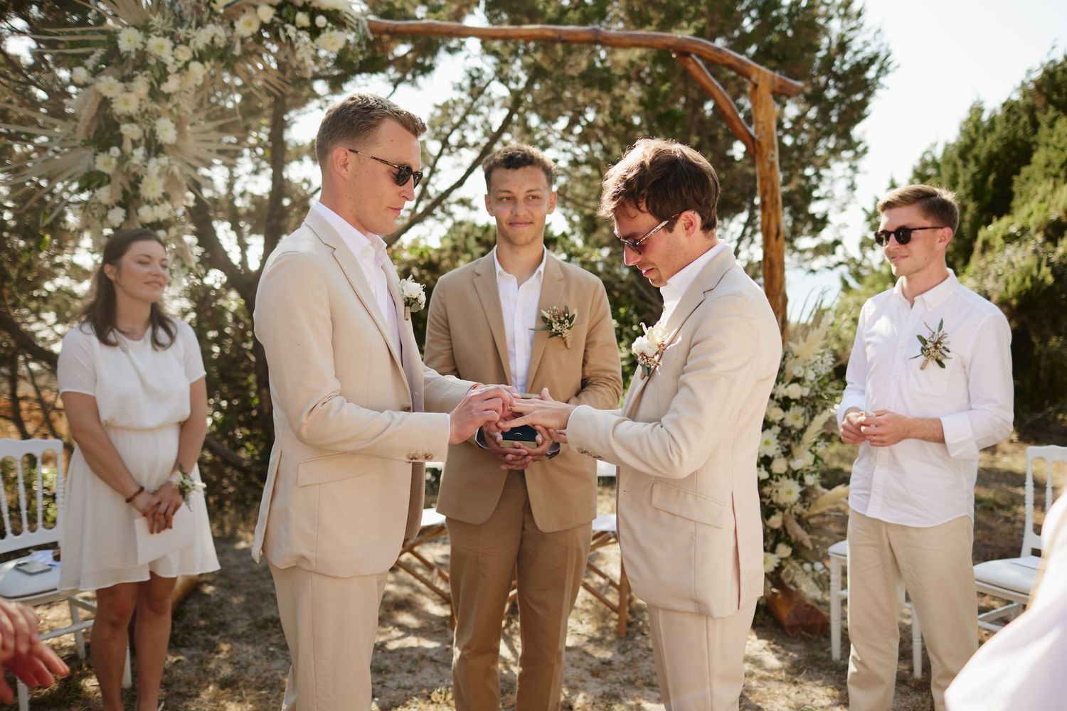 Two grooms exchange rings under a floral arch at their outdoor wedding ceremony.