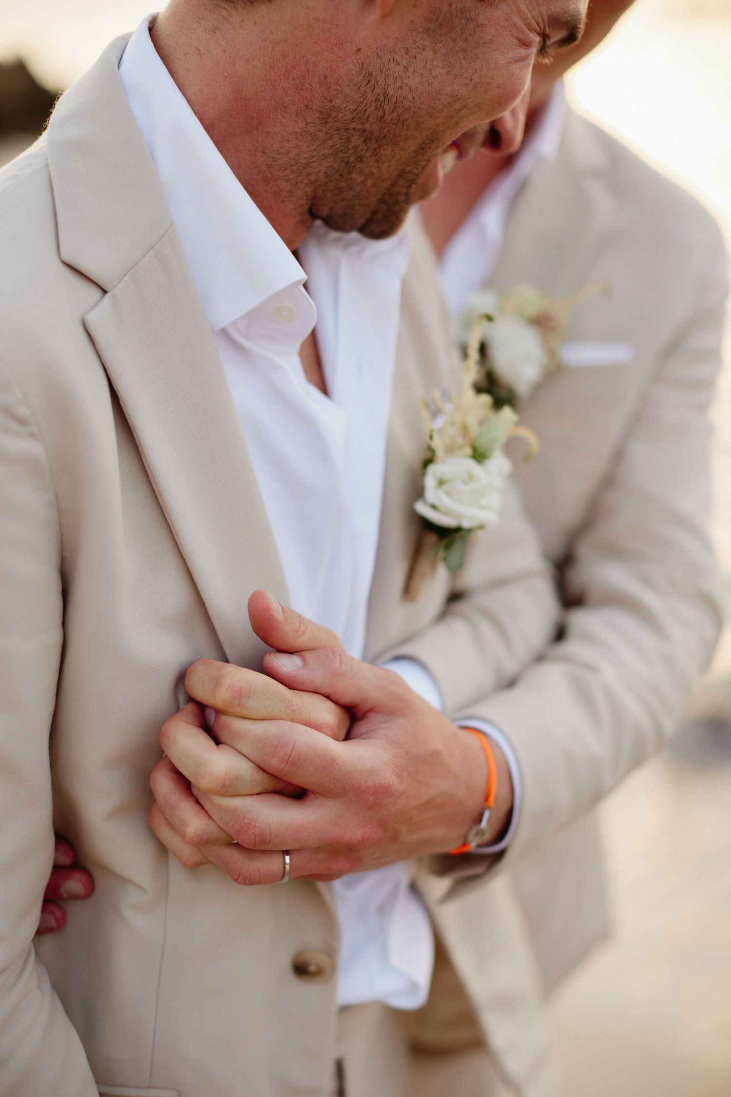 Close-up of two grooms holding hands, one wearing a wedding ring and floral boutonniere.
