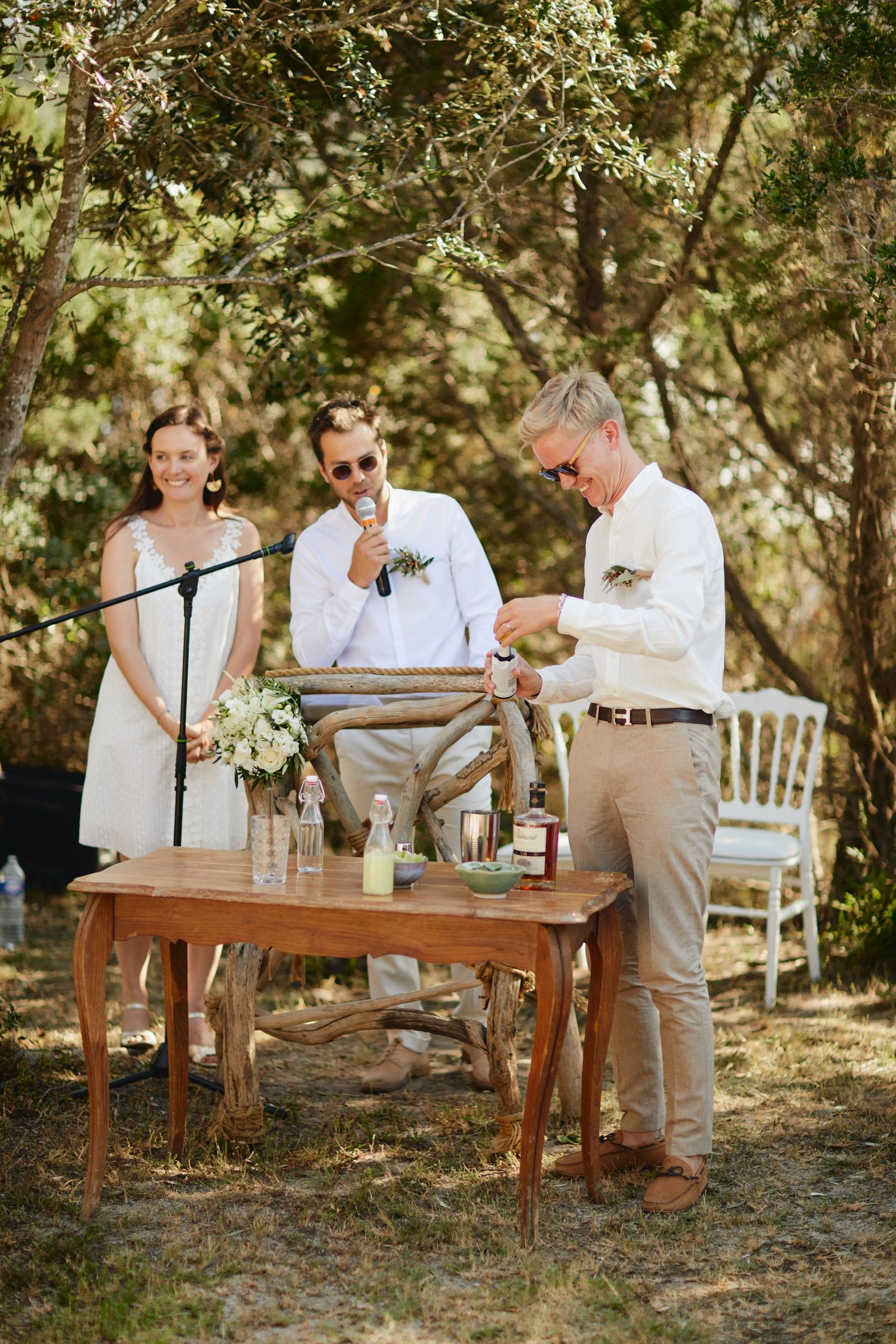 Groomsman opens champagne bottle at rustic wooden table during outdoor wedding ceremony in forest.
