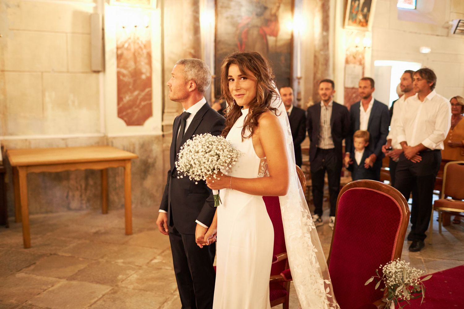 Bride in elegant white gown and veil walking up church aisle holding baby's breath bouquet during ceremony.