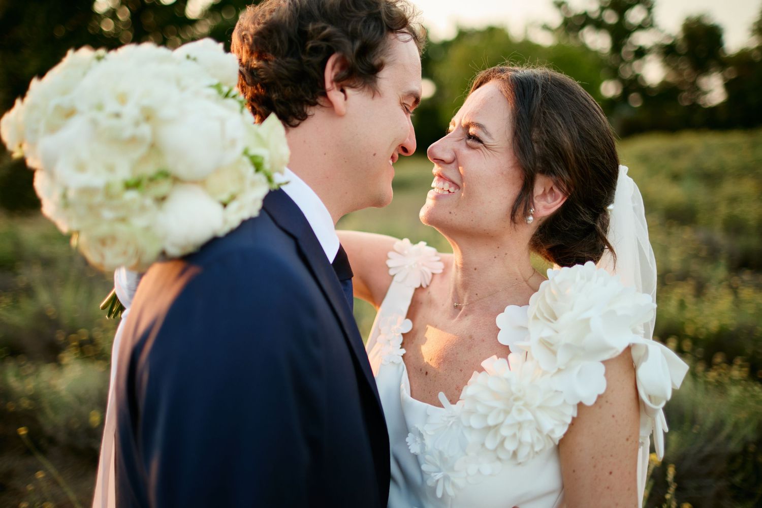 Smiling bride and groom share intimate moment with white floral bouquet at golden hour.