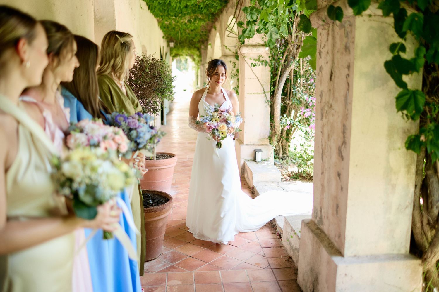 Bride in white gown walking toward bridesmaids holding colorful bouquets in a vine-covered corridor.