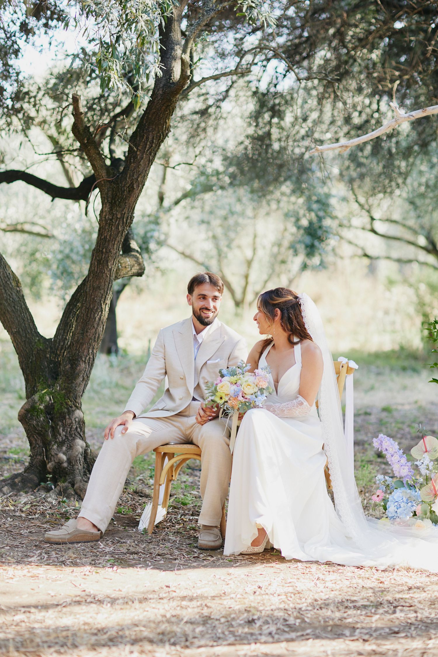 Smiling bride and groom share a tender moment seated together at outdoor woodland wedding.