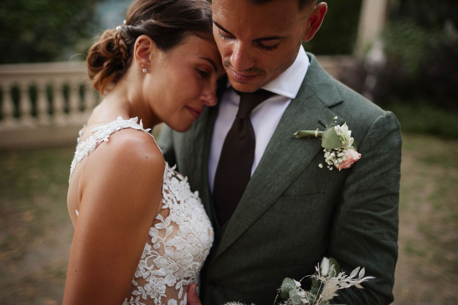 Close-up of bride leaning on grooms chest in green linen suit with floral boutonniere at outdoor wedding.
