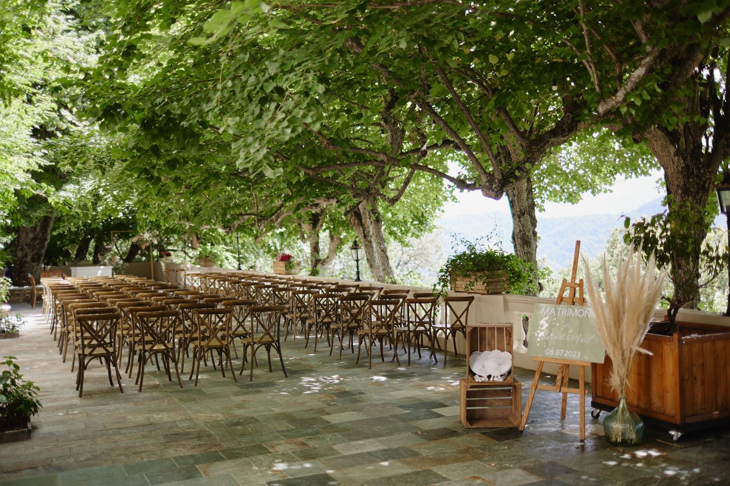 Rustic outdoor wedding ceremony setup with wooden chairs under lush green trees at a scenic venue.