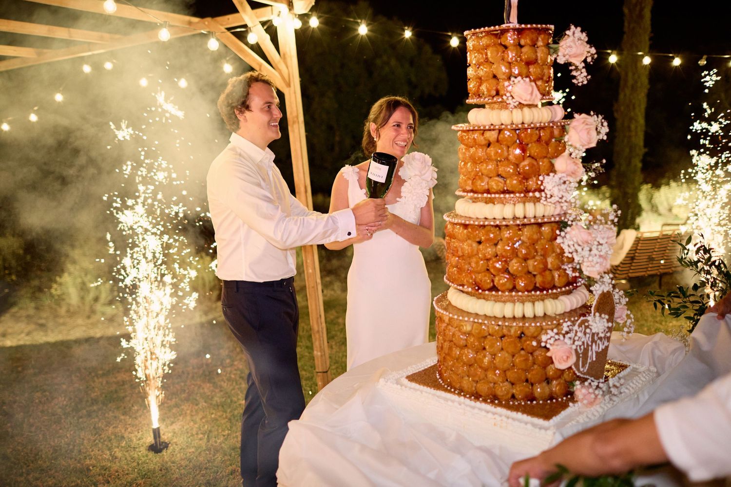 Bride and groom toasting beside elegant tiered croquembouche wedding cake with sparklers outdoors.