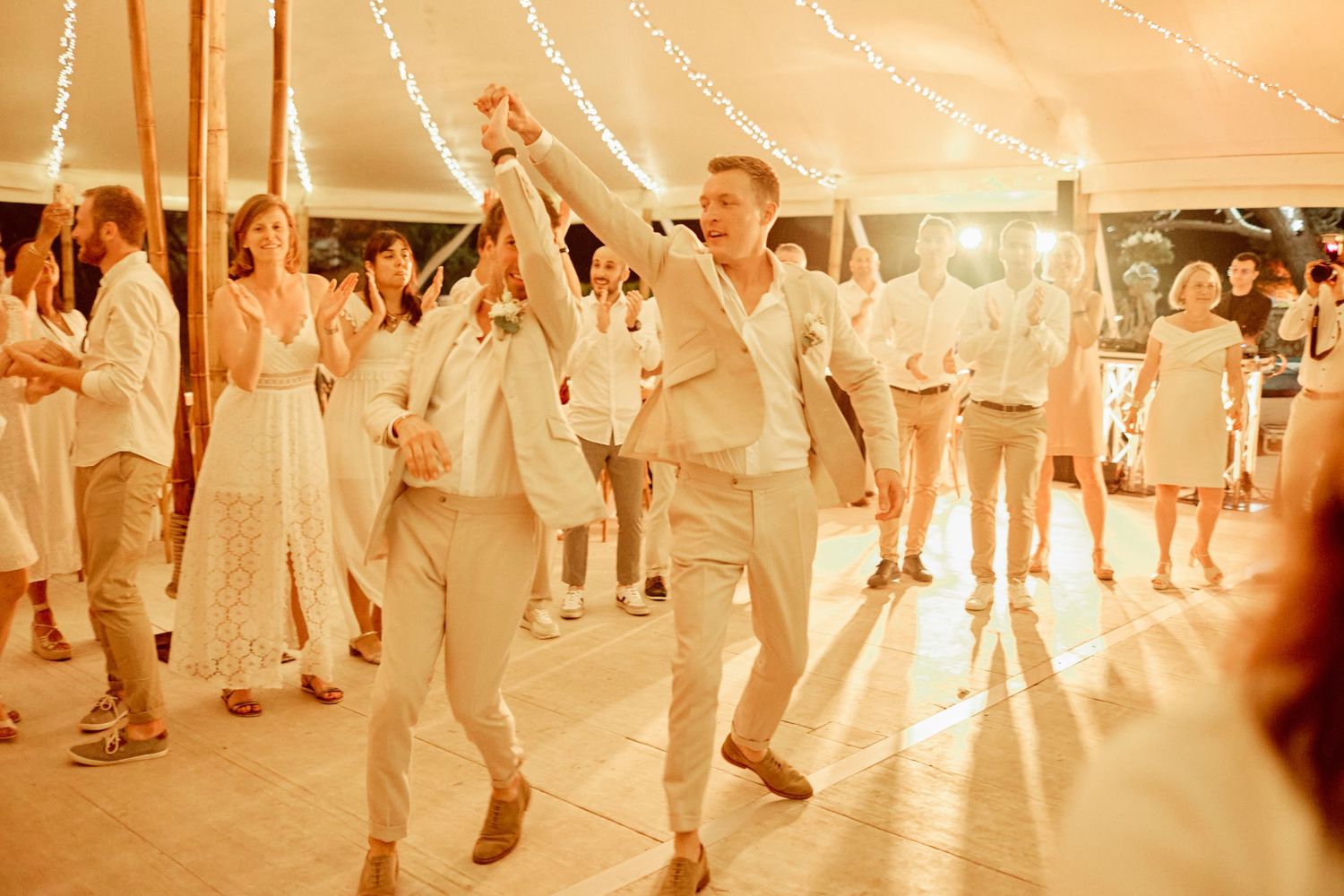 Grooms dance joyfully with wedding guests under fairy lights in a warm reception tent.