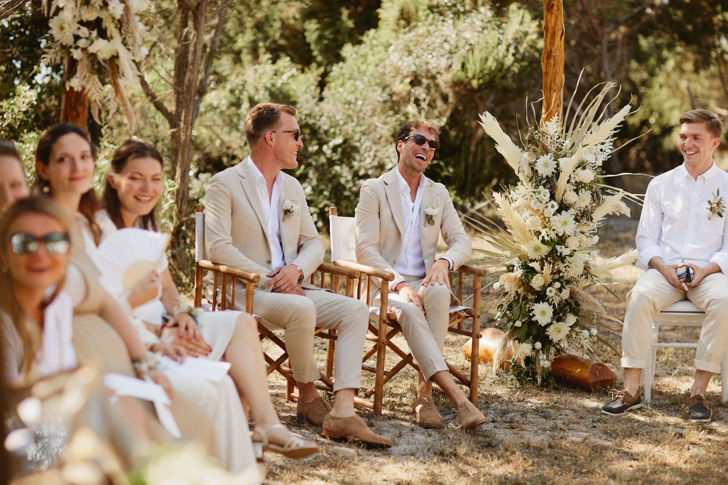 Two grooms in tan suits laughing with guests at sunny outdoor wedding.