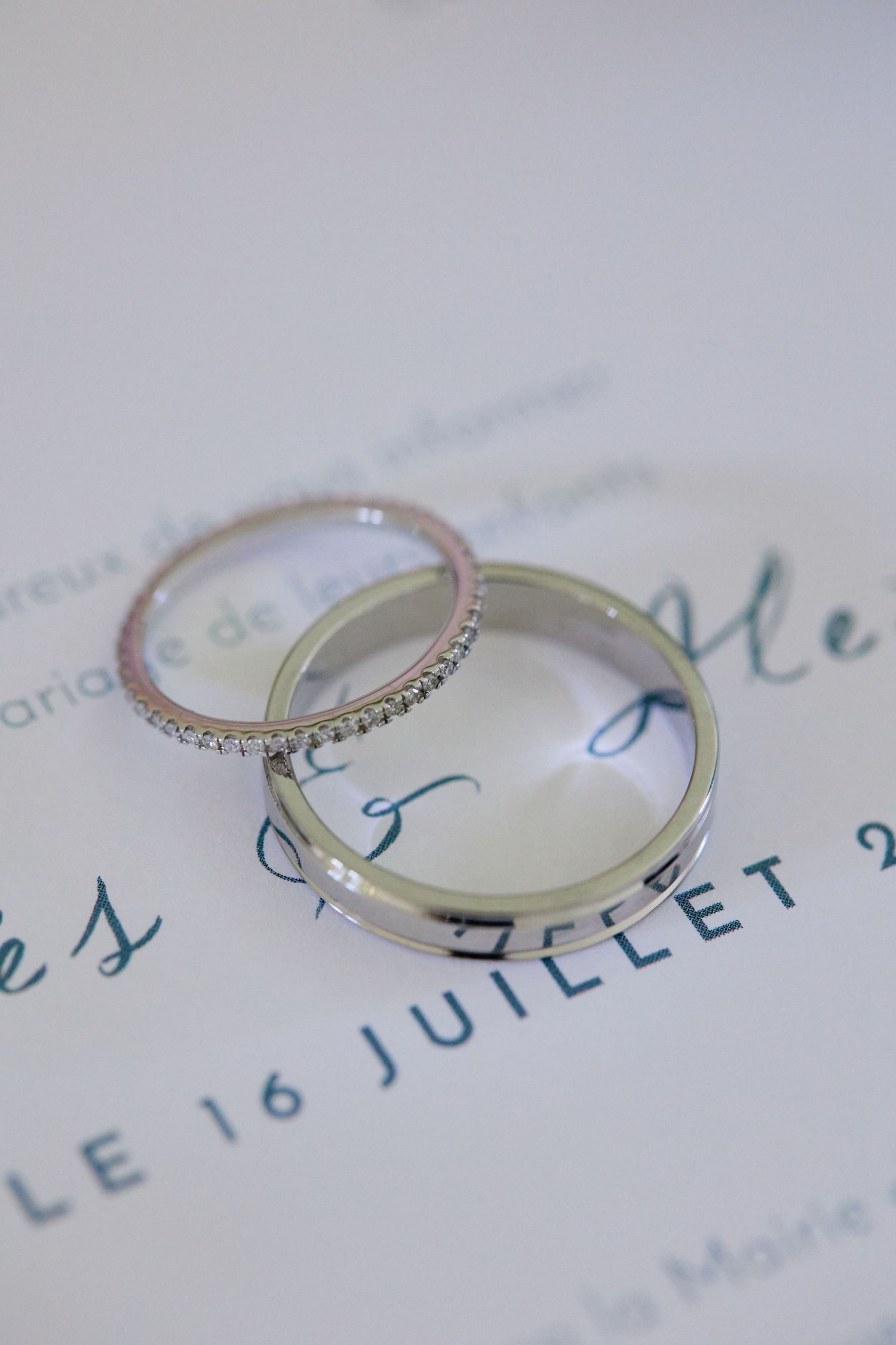 Two silver wedding bands resting on a wedding invitation reading Le 16 Juillet 2023.
