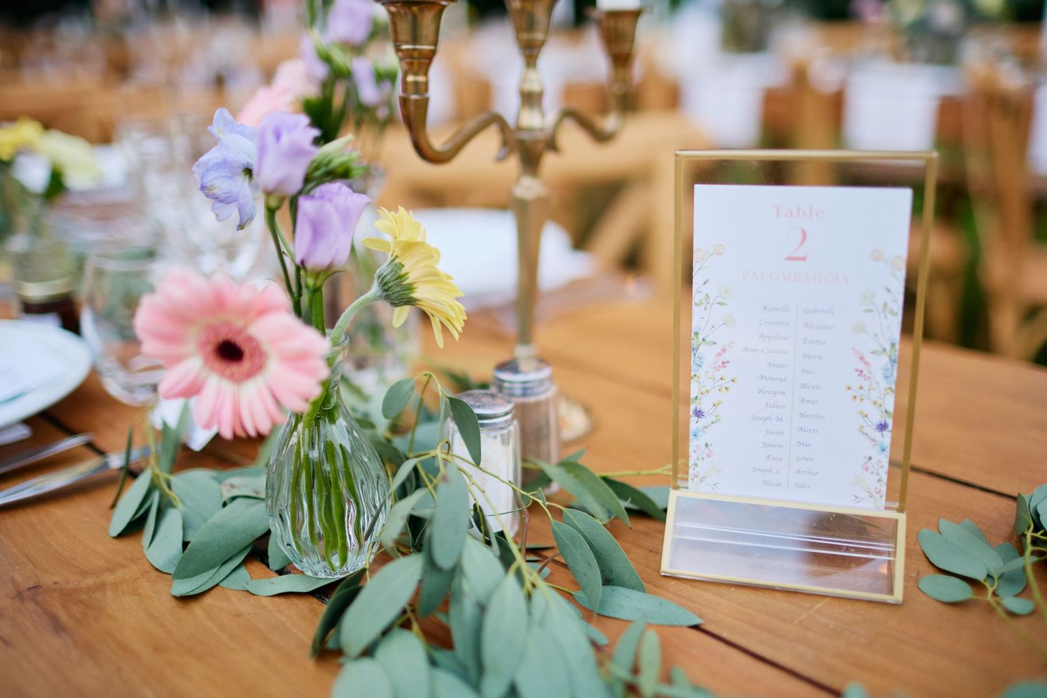 Wedding table detail with gerbera daisy florals, eucalyptus garland, and framed table number card on wood.