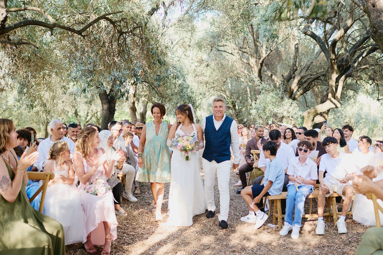 Bride walks down outdoor aisle flanked by parents among olive trees at rustic wedding ceremony.