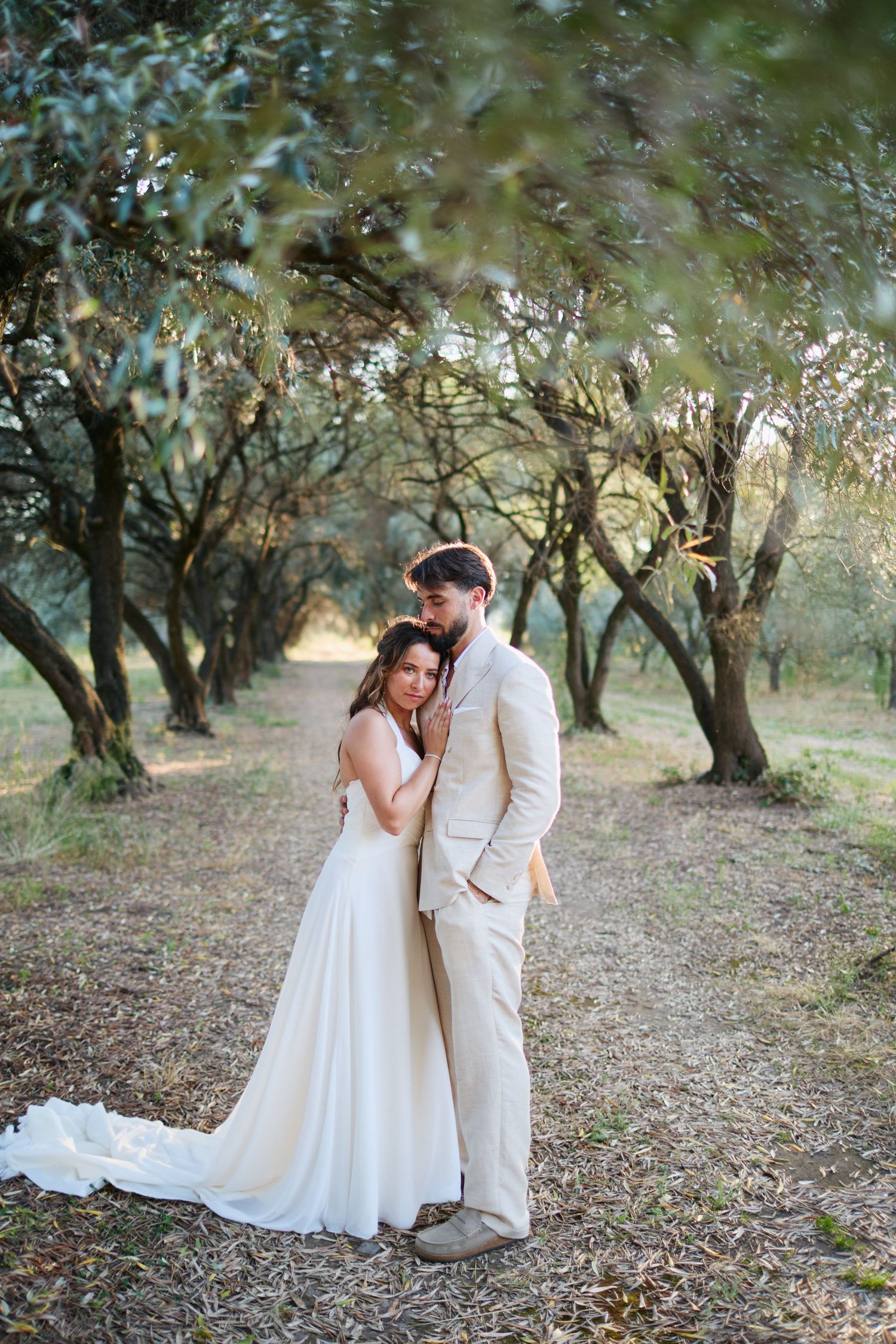 Wedding couple sharing a tender embrace on a tree-lined path during golden hour in an olive grove.