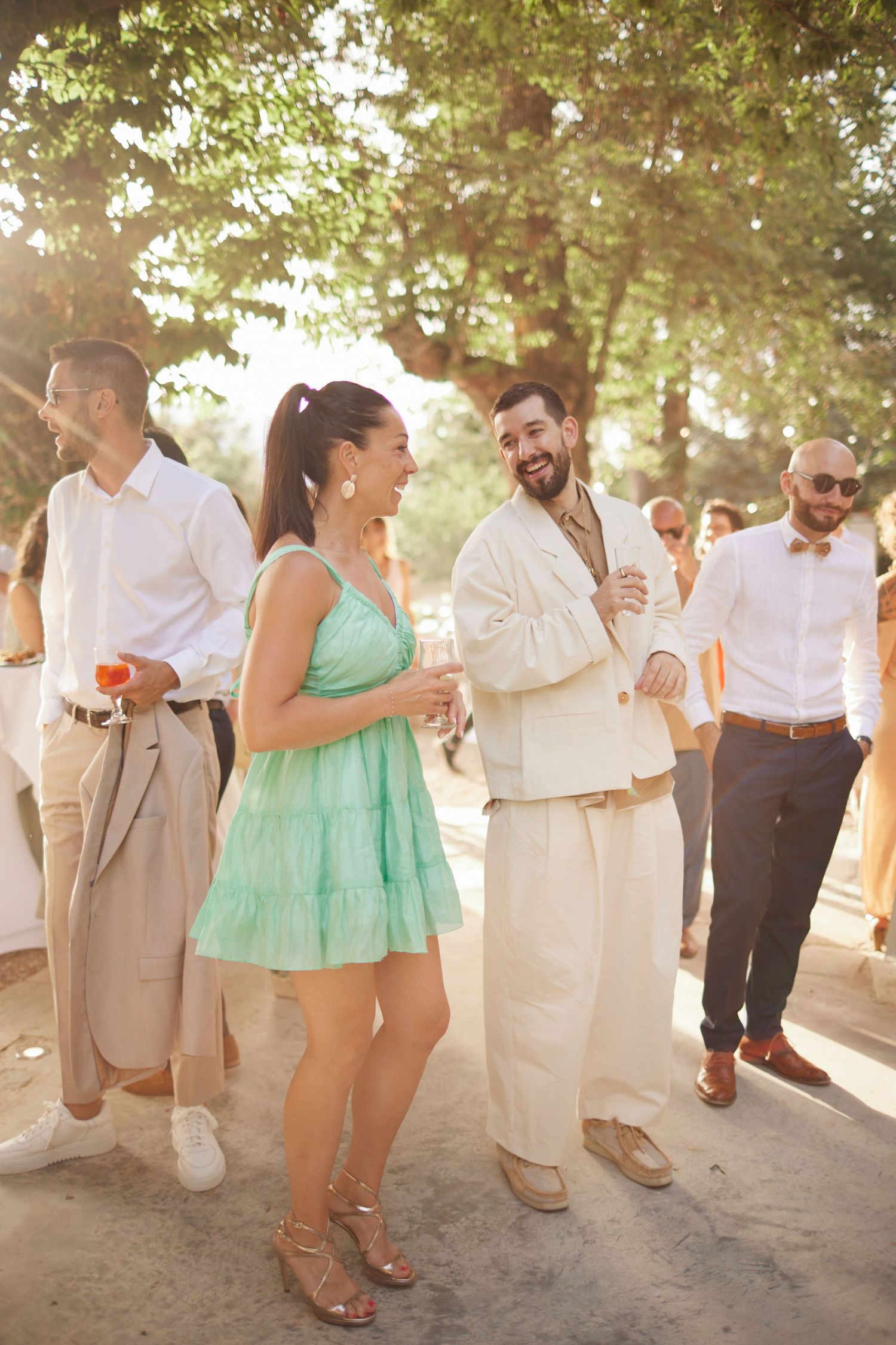 Wedding guests in summer attire laughing and socializing outdoors in golden sunlight.
