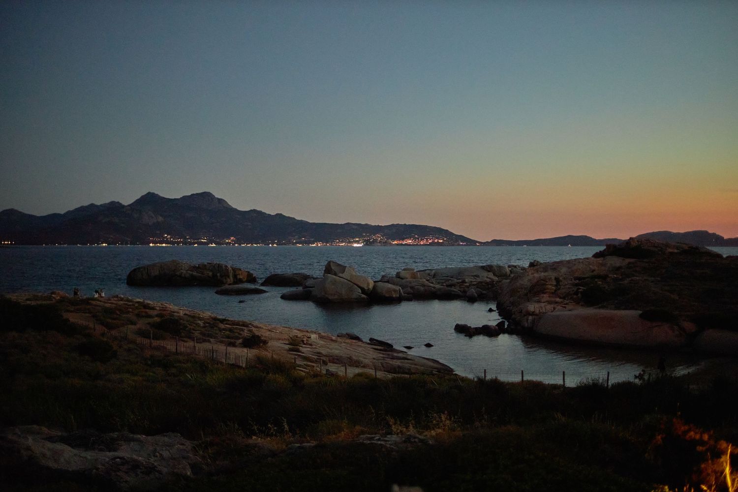 Scenic coastal sunset view with rocky shoreline, calm bay, and distant mountain silhouette.