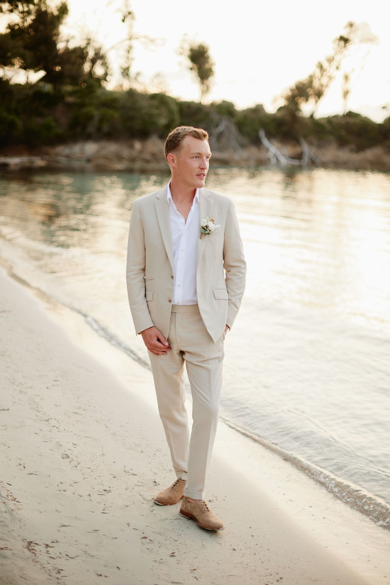 Groom in beige suit with boutonniere walking along a serene beach shoreline at sunset.