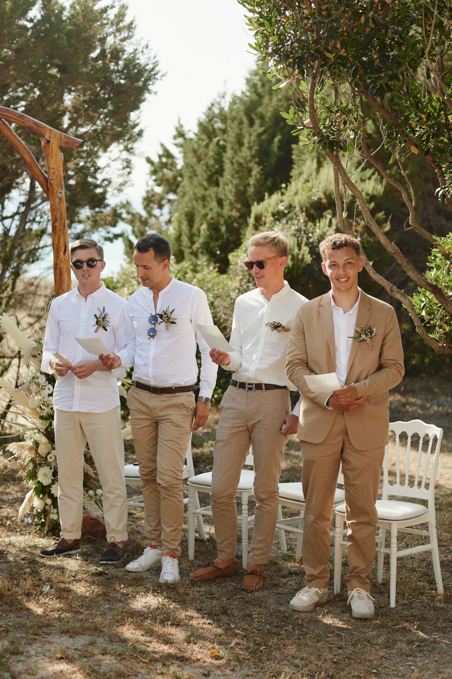 Four groomsmen in beige suits and white shirts stand outdoors at a wedding ceremony among trees.