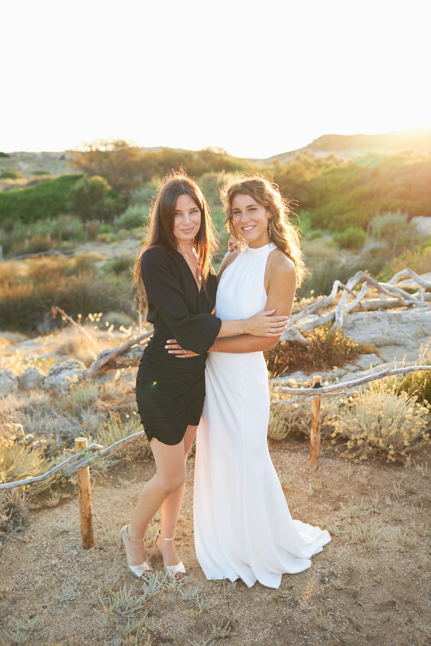 Bride in elegant white gown posing with guest in black dress at sunset outdoor wedding.