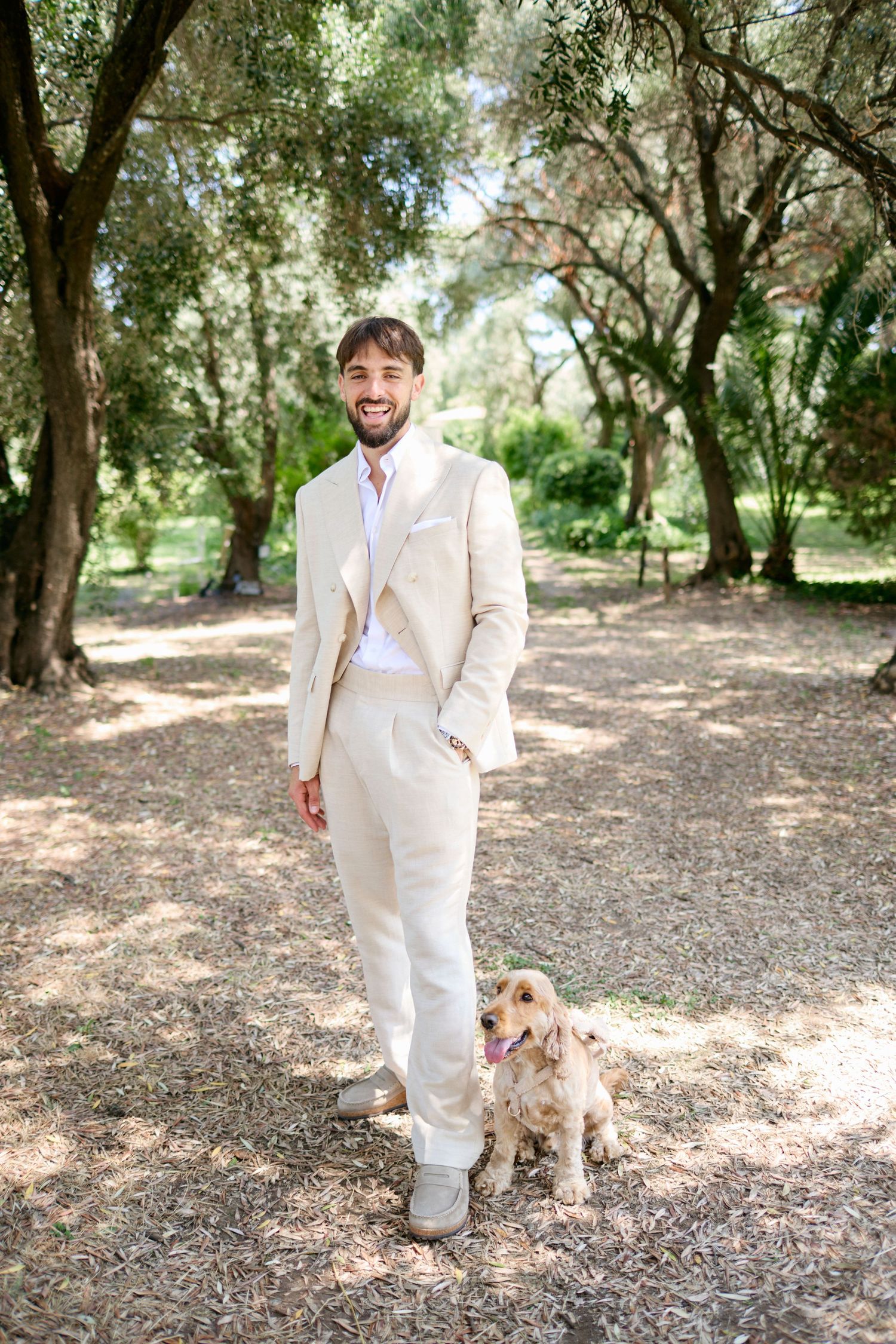 Smiling groom in beige linen suit standing with a golden cocker spaniel in a shaded garden.