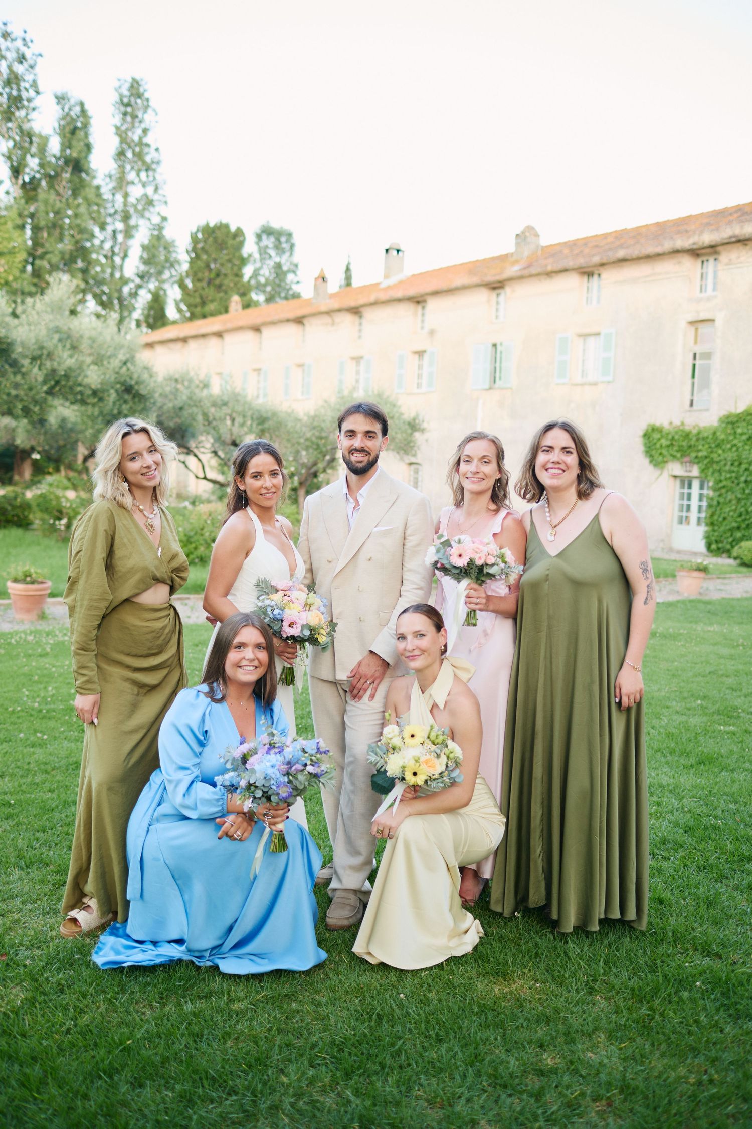 Wedding party group photo with bride, groom, and guests in colorful attire on lawn at French chateau.