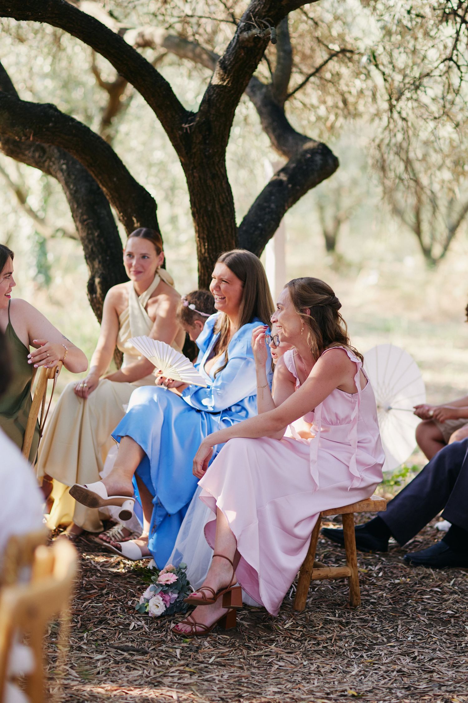 Wedding guests in pastel dresses fan themselves while seated under olive trees at outdoor ceremony.