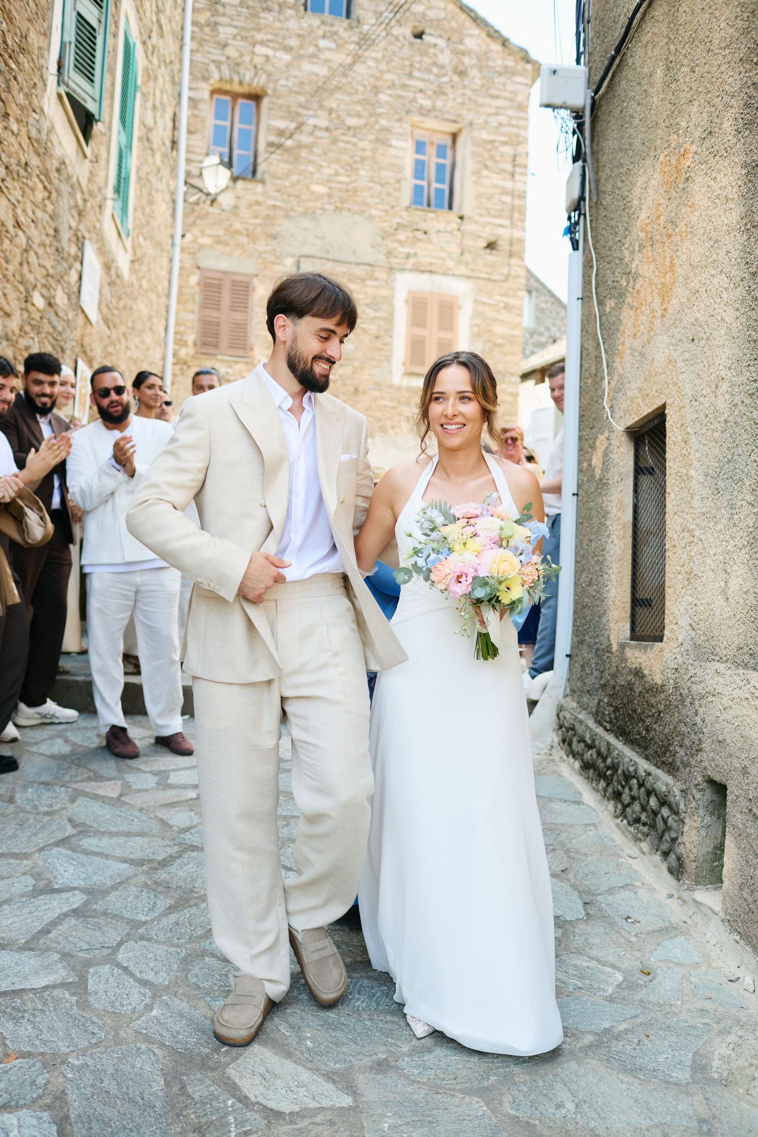 Newlyweds walk cobblestone street in historic village as guests celebrate around them.