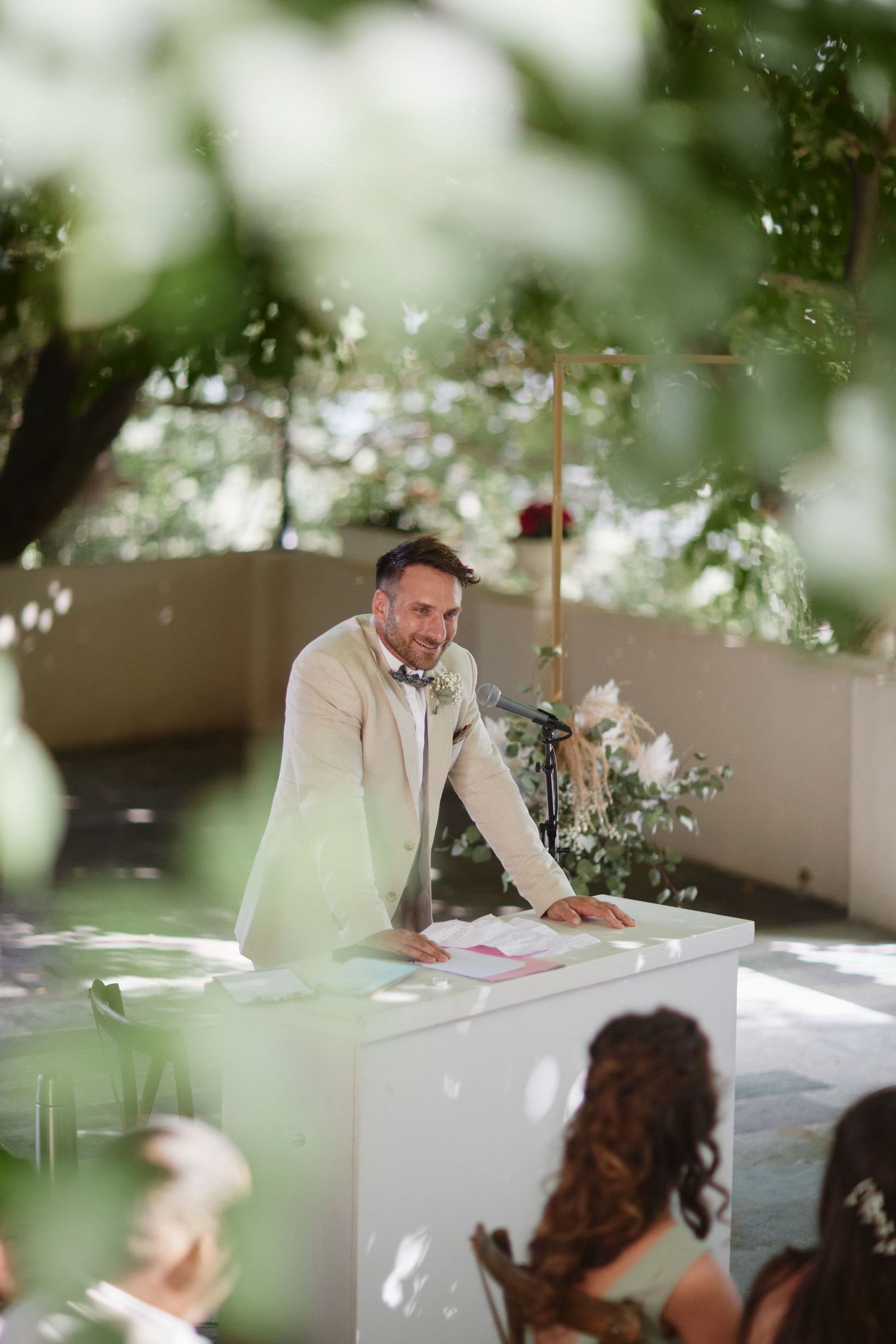 Smiling groom in cream blazer and bow tie officiating outdoor wedding ceremony beneath leafy green trees.