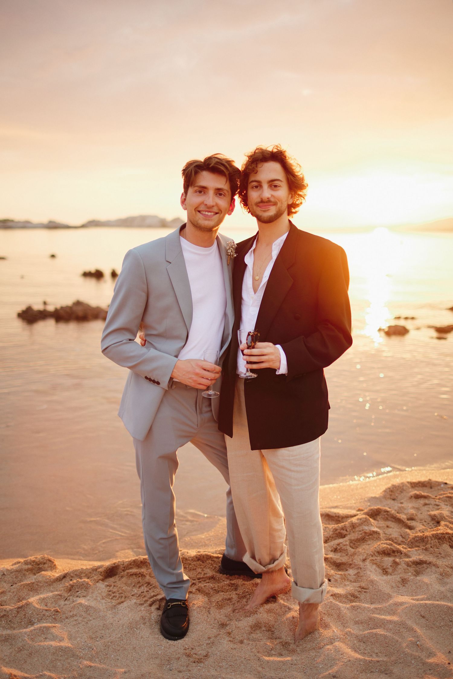 Two stylish male wedding guests posing together on a sandy beach during a stunning sunset.
