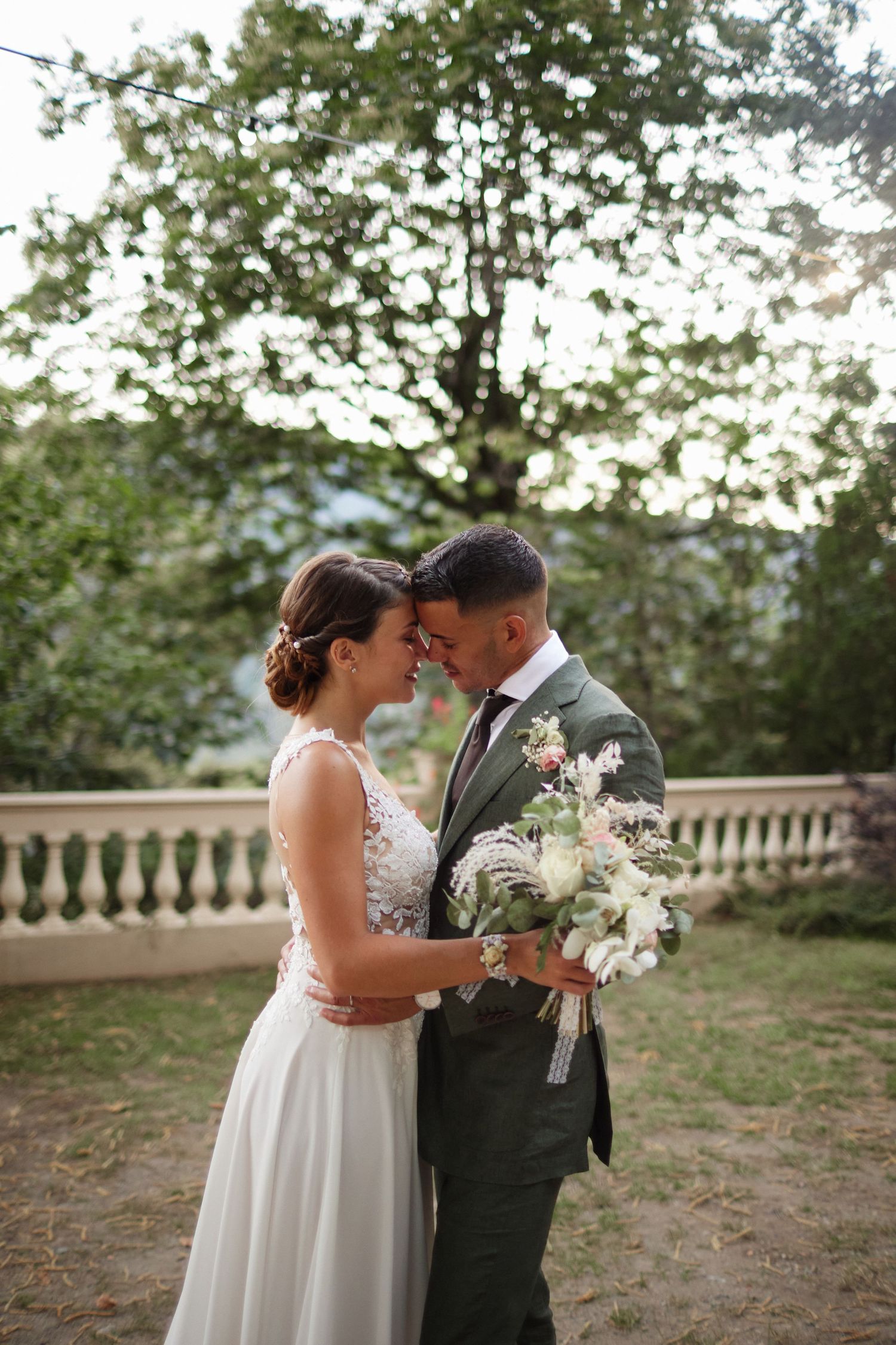 Bride and groom share an intimate forehead touch in sunlit garden, groom holding elegant white floral bouquet.