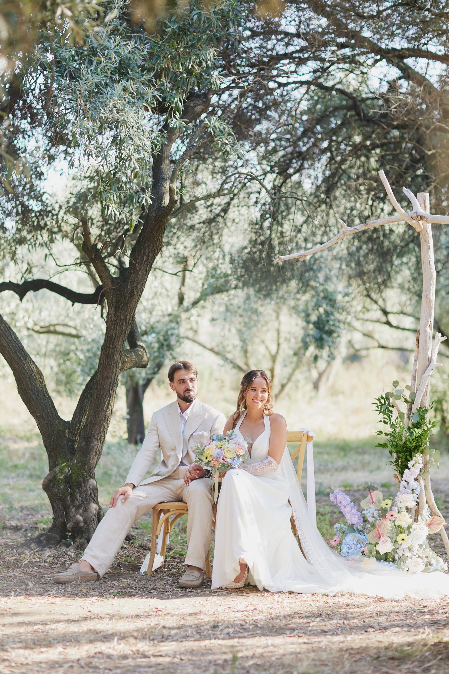 Bride and groom sit together under olive tree at rustic outdoor wedding ceremony altar.