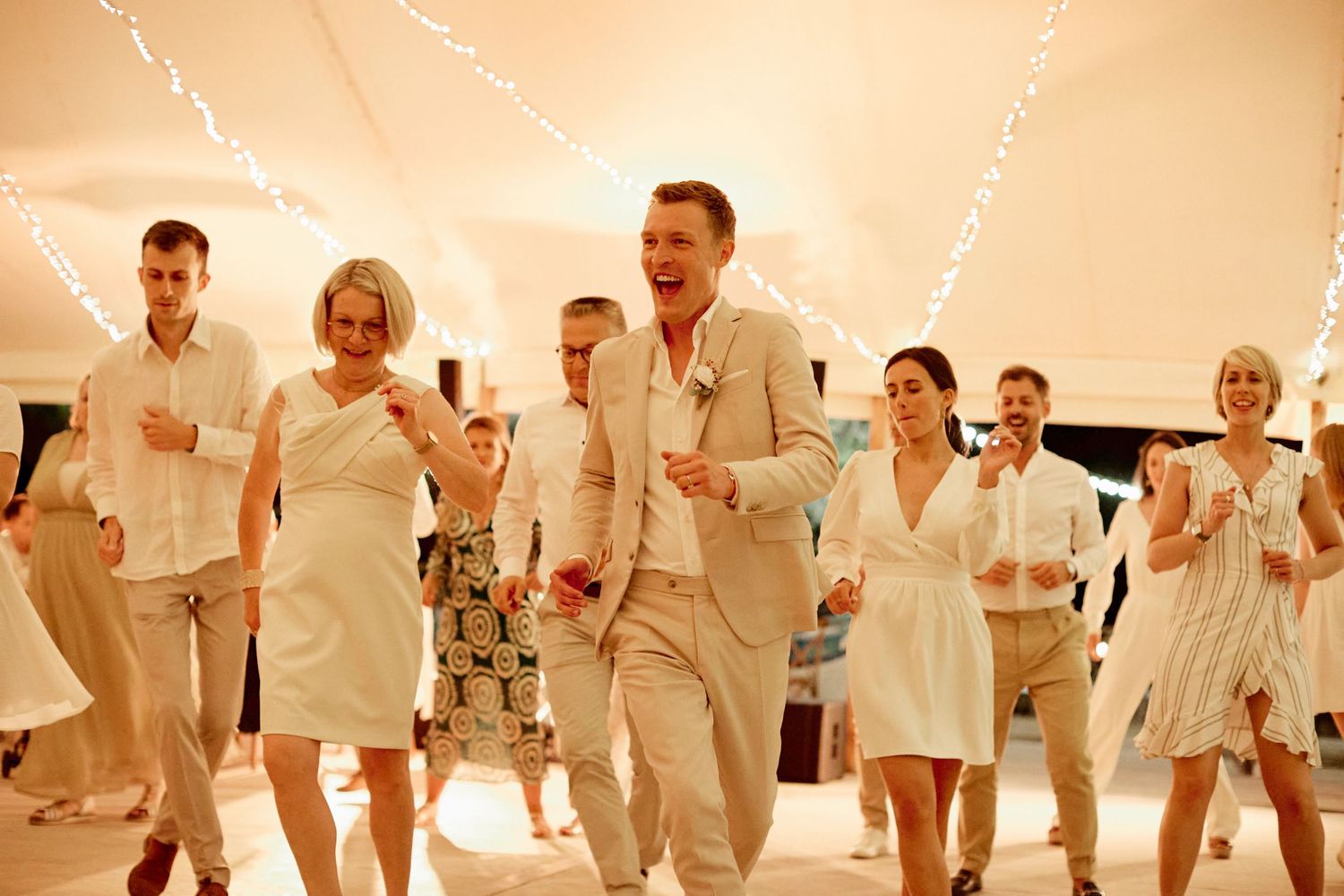Wedding guests in white and beige attire dancing joyfully under fairy lights at a beach reception.