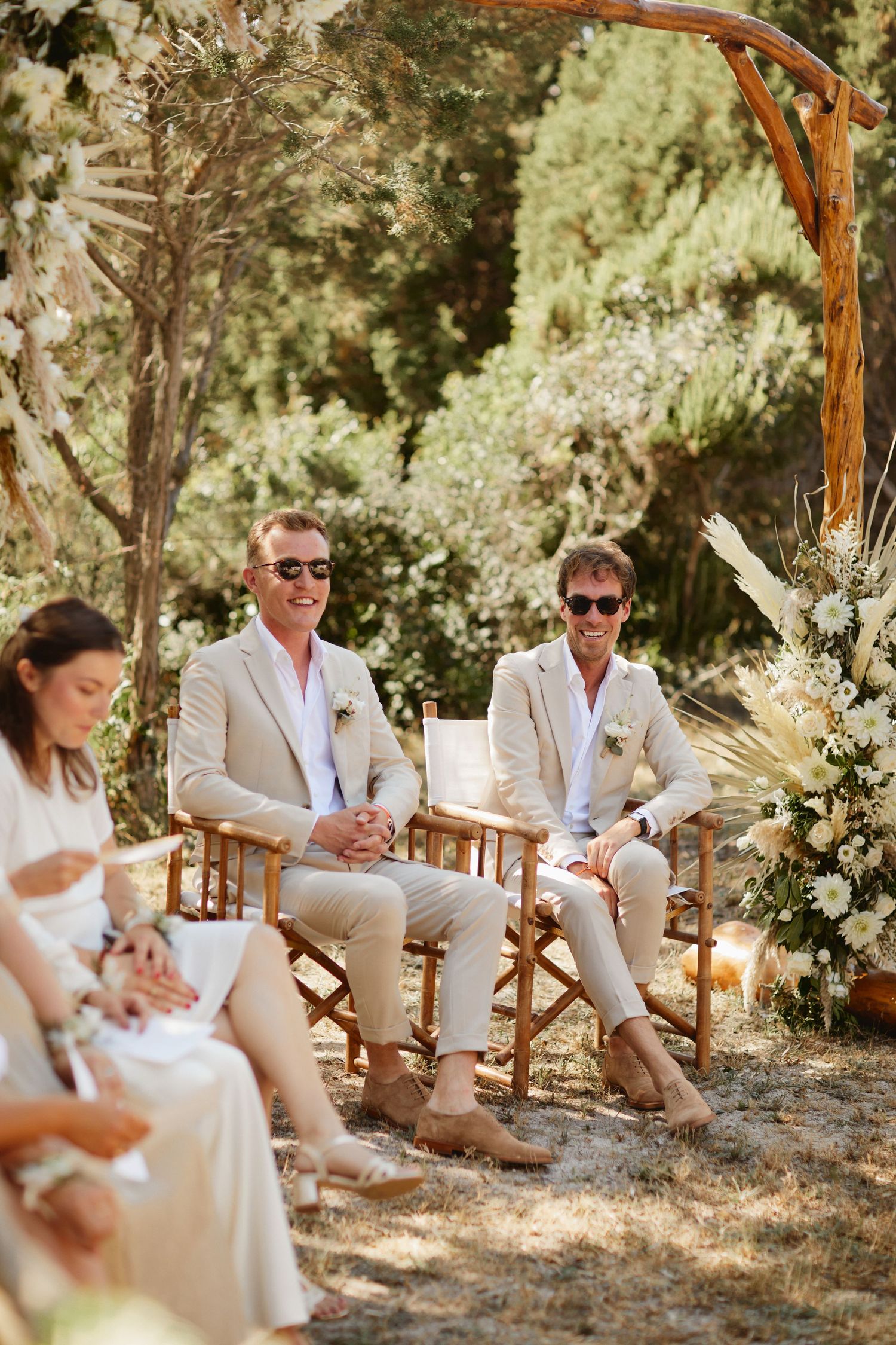 Two grooms in beige suits smiling under wooden arch at outdoor ceremony.