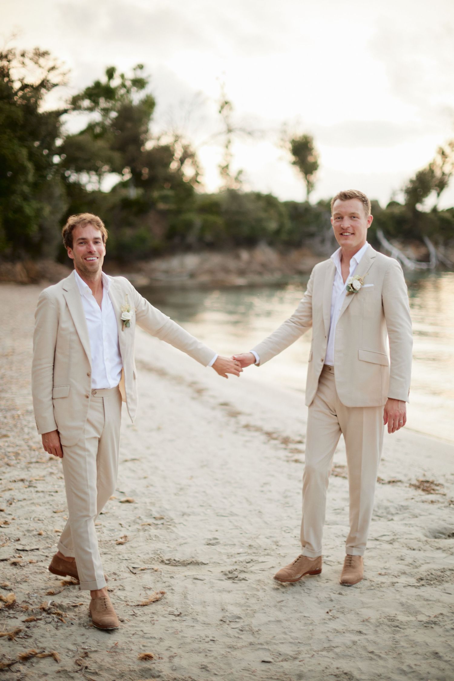 Two grooms in cream linen suits smile and hold hands while walking on a tropical beach.