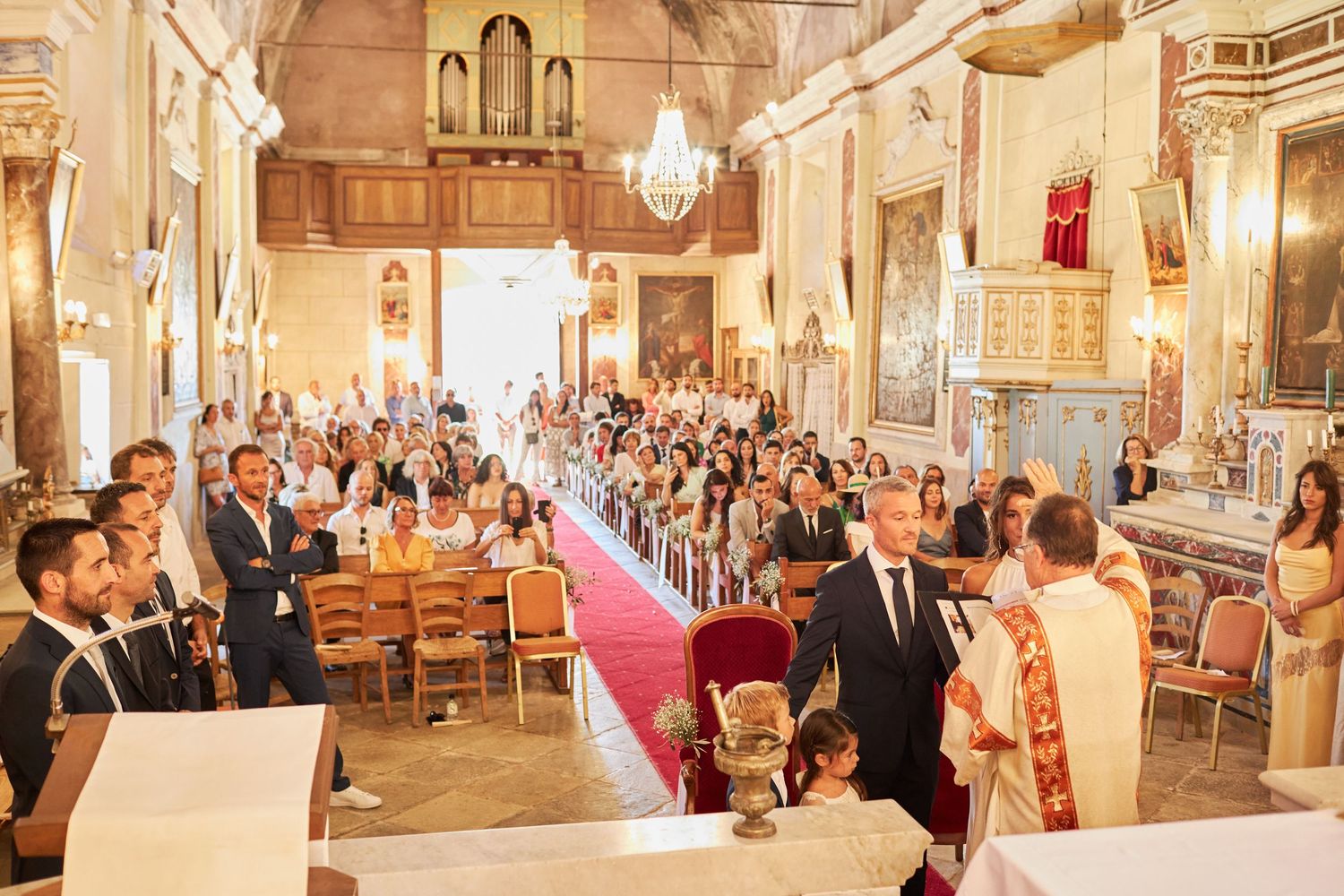 Wide view of packed church wedding ceremony with guests seated in pews along red carpet aisle.