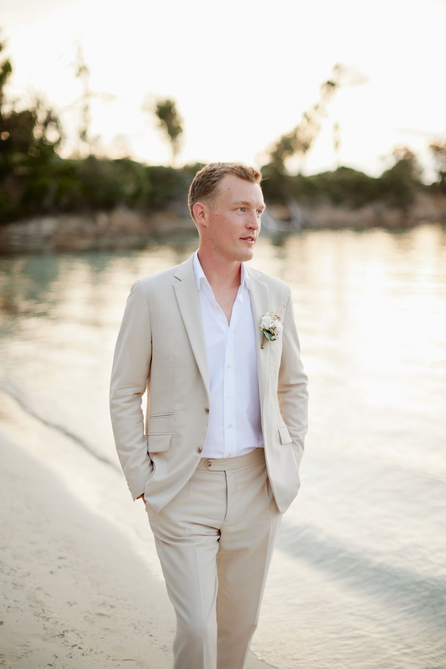Handsome groom in tan linen suit with boutonniere posing on a tropical beach at dusk.