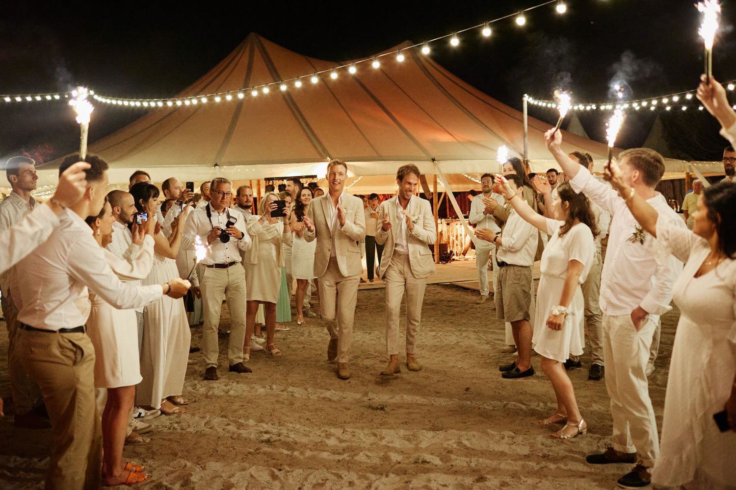 Wedding couple and guests celebrating with sparklers outside a glowing sailcloth tent on a sandy beach at night.
