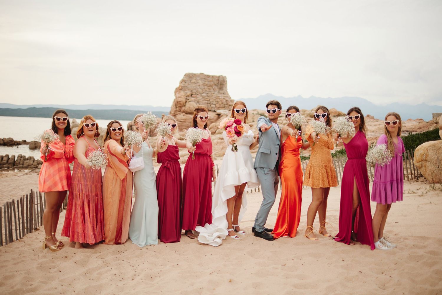 Colorful bridesmaids in vibrant dresses posing with bride and groom on sandy beach wedding.