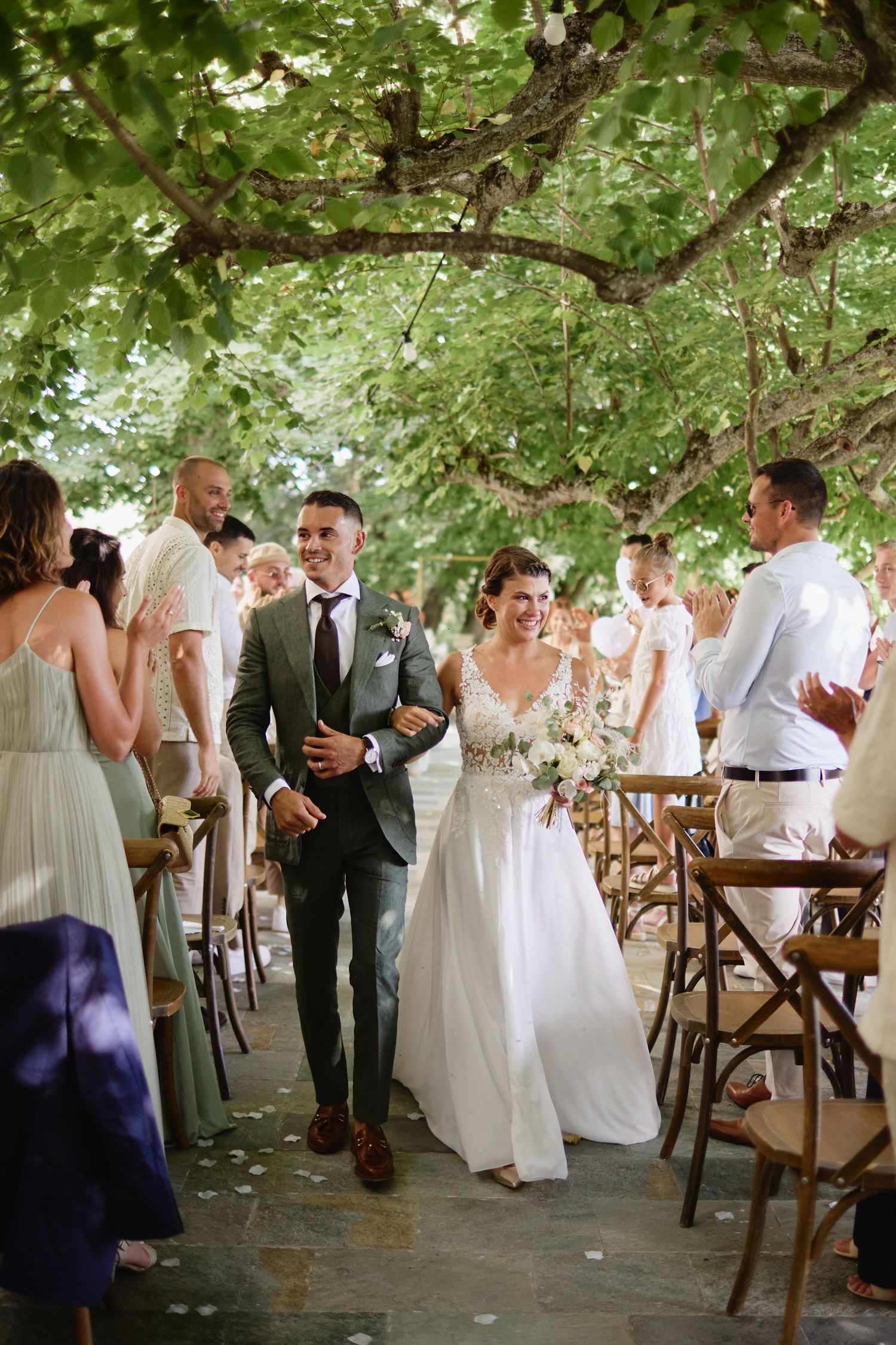 Radiant newlyweds walking down the aisle together after outdoor wedding ceremony under green tree canopy.