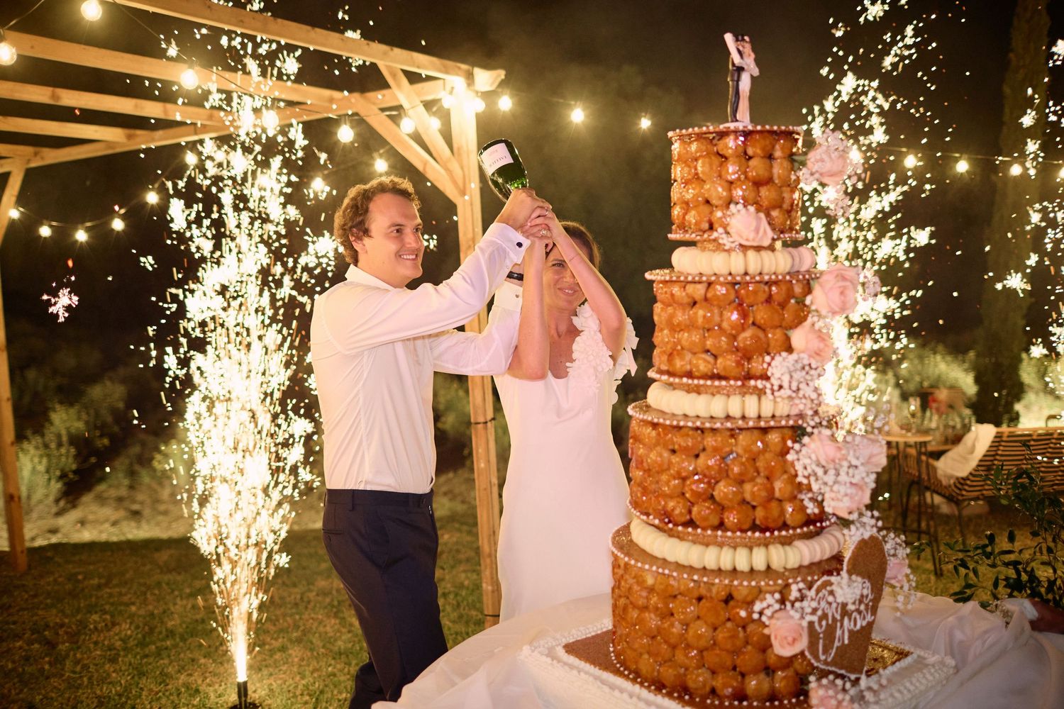 Couple celebrating with champagne beside tall croquembouche wedding cake with sparklers at night.